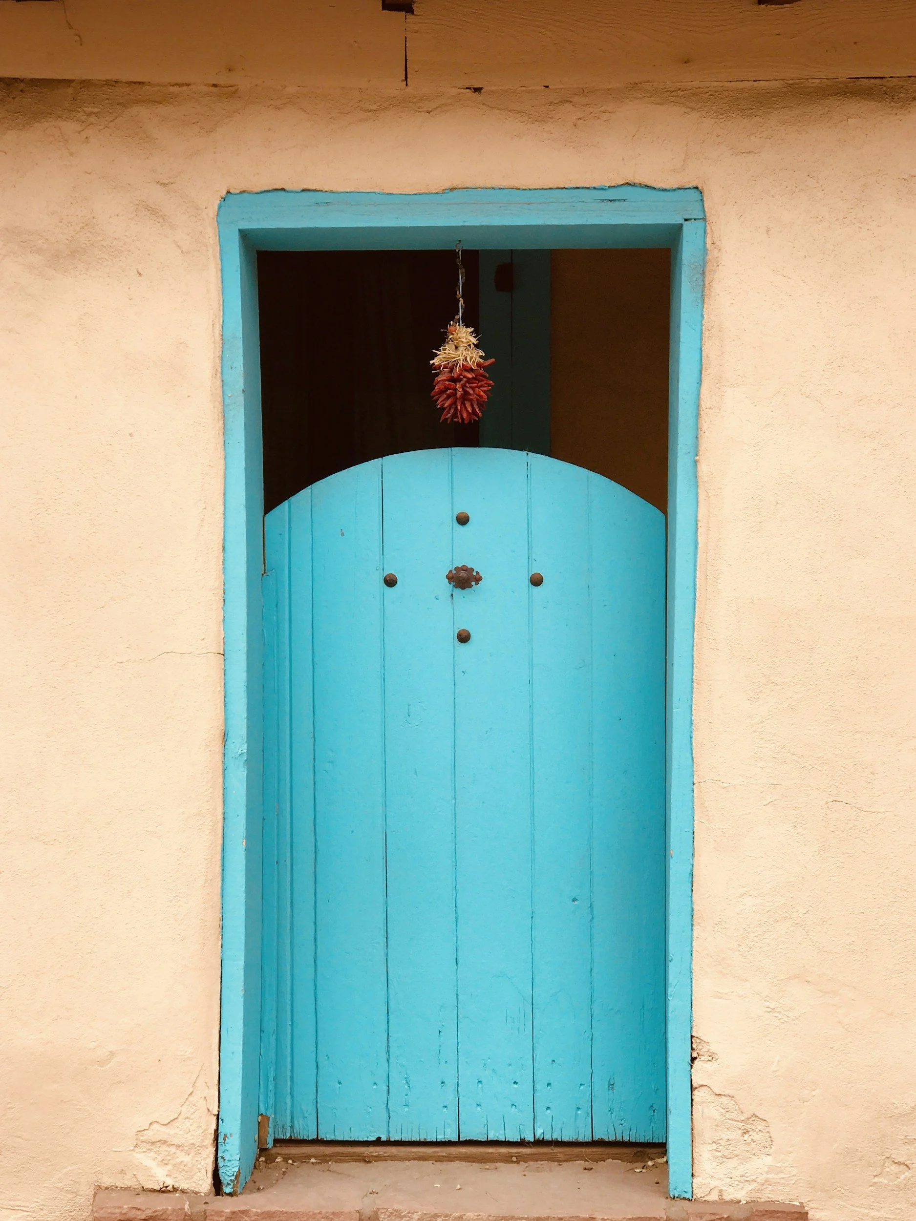Blue wooden door with a half-circle top, set in a beige stucco wall, decorated with hanging chili peppers above.