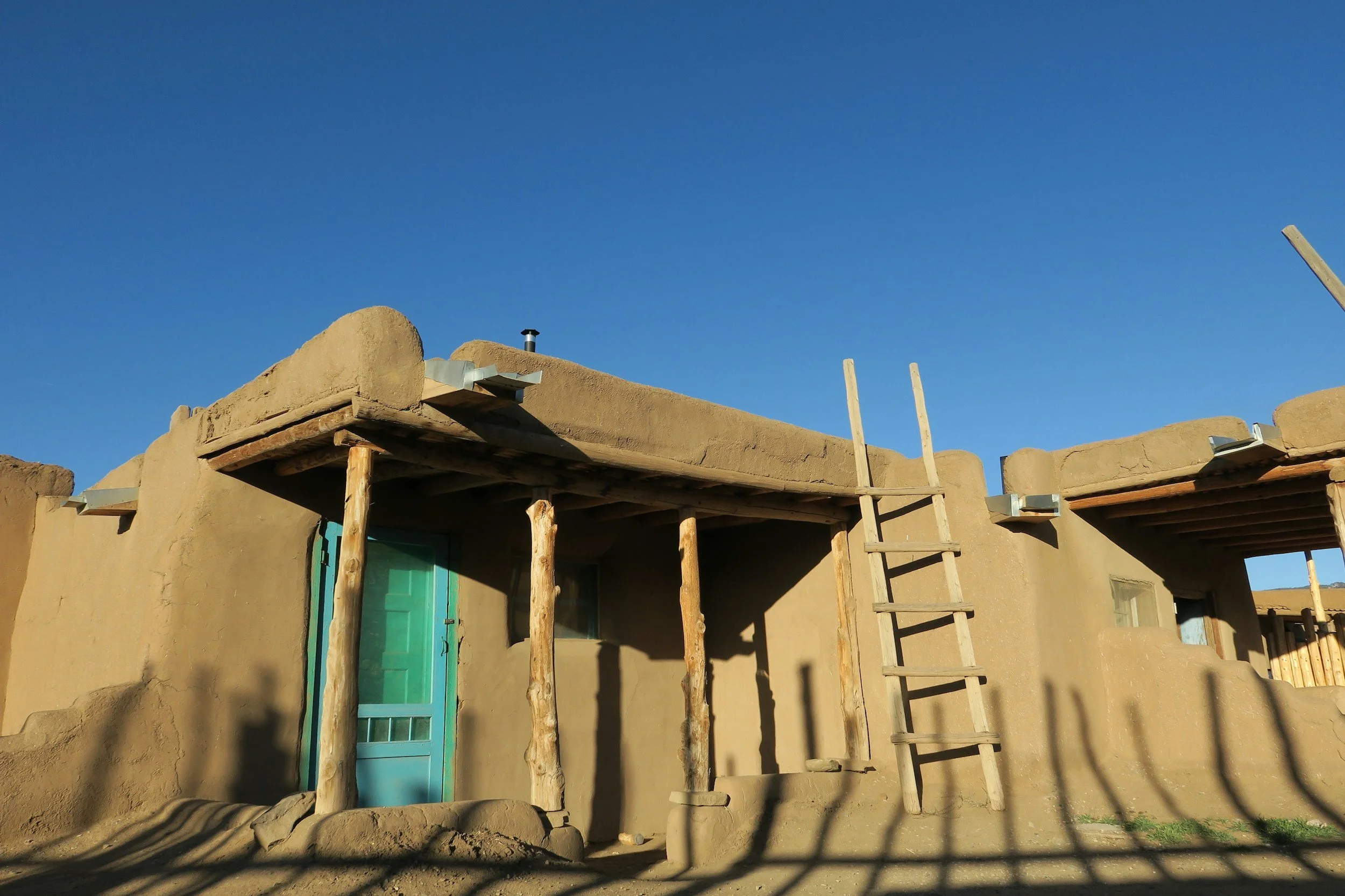 A traditional adobe-style house under construction with wooden supports, ladder, and blue door, set against a clear blue sky.