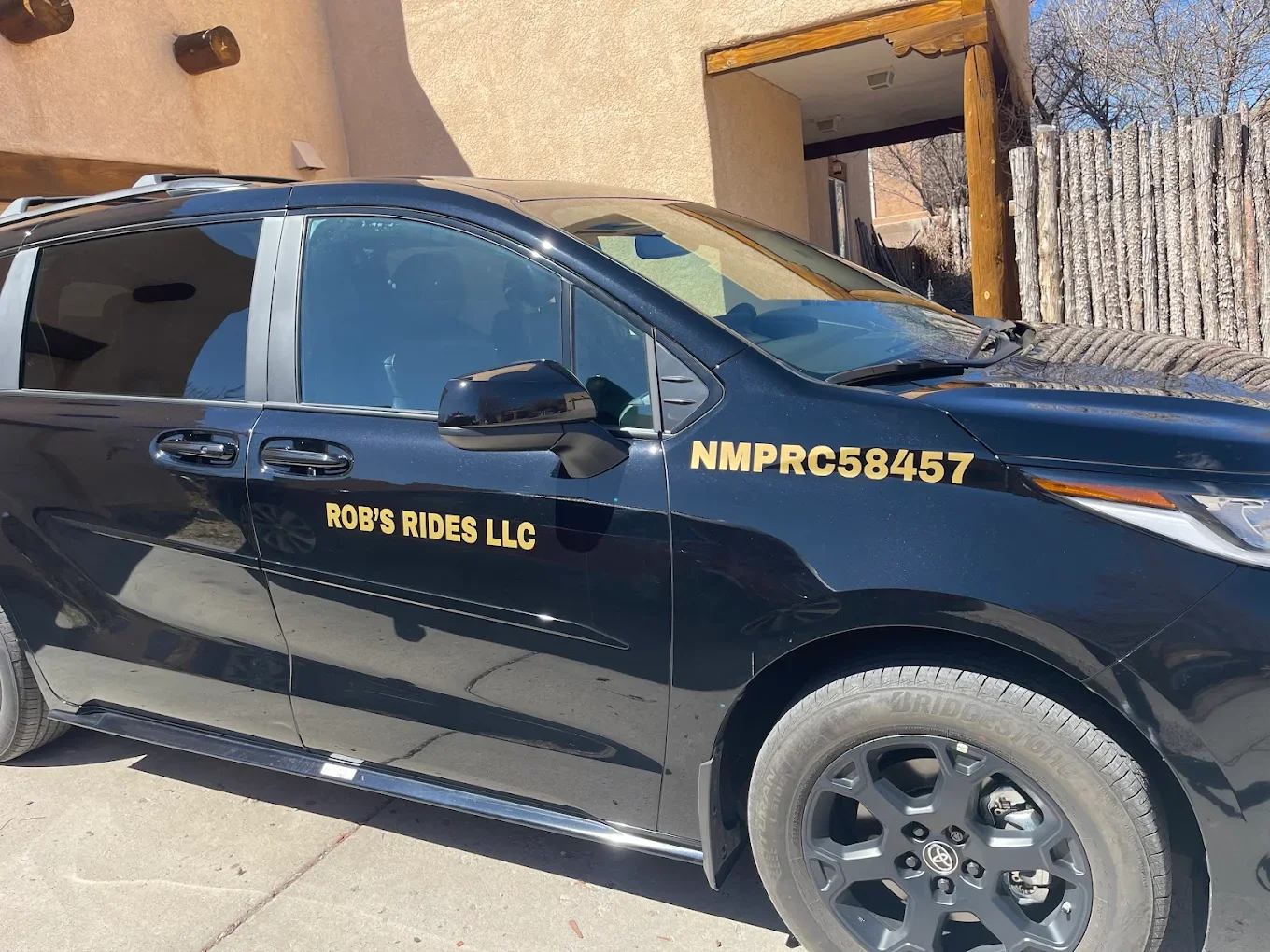 Black vehicle with gold lettering reading 'ROB'S RIDES LLC' and a license number 'NMPRC58457' parked on a concrete driveway next to a building with a beige wall and a wooden fence.