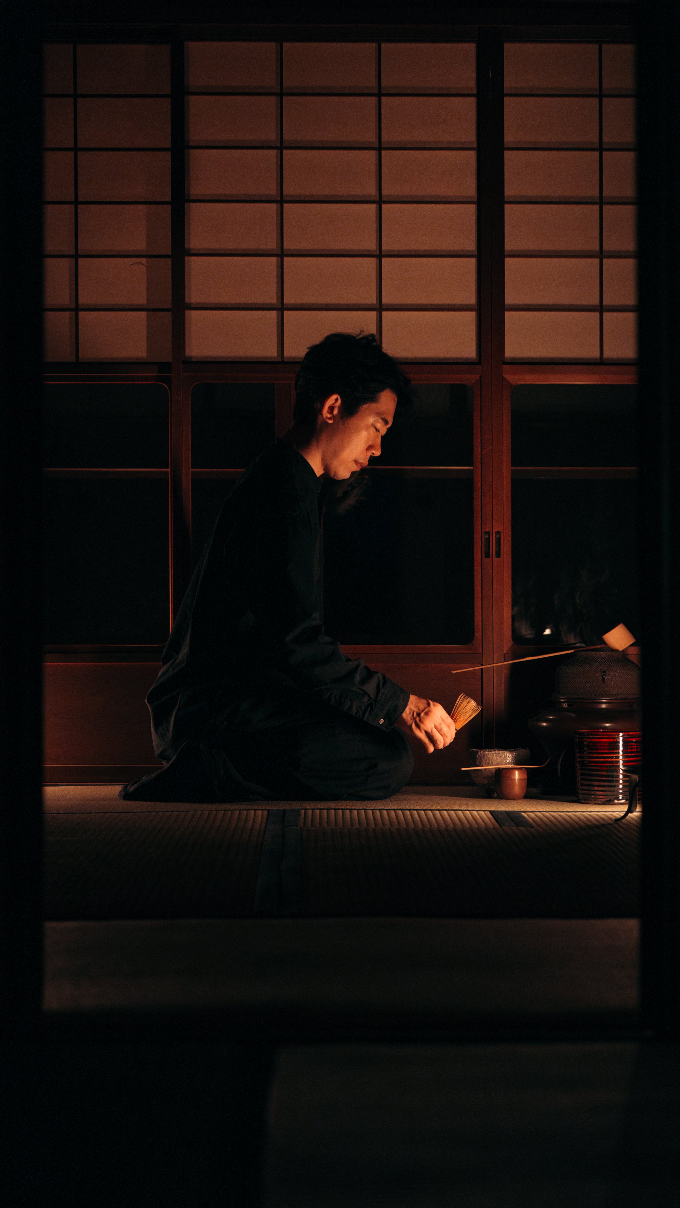 A man dressed in black sitting on a tatami mat floor in a Japanese-style room, preparing tea with traditional tea utensils, illuminated by soft warm light.