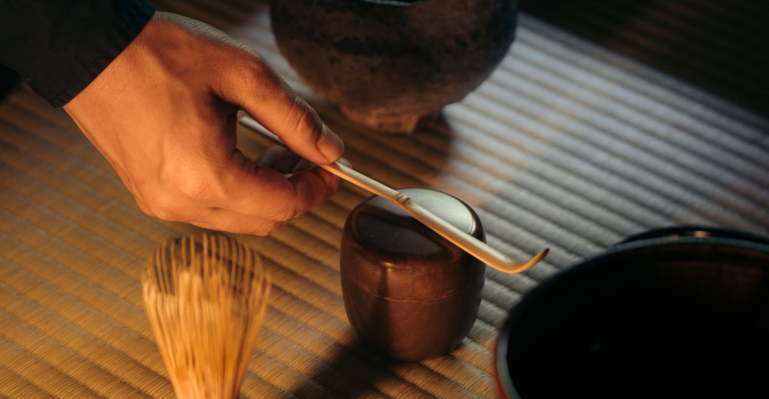 A person’s hand holding a matchstick over a small ceramic tea cup on a tatami mat.