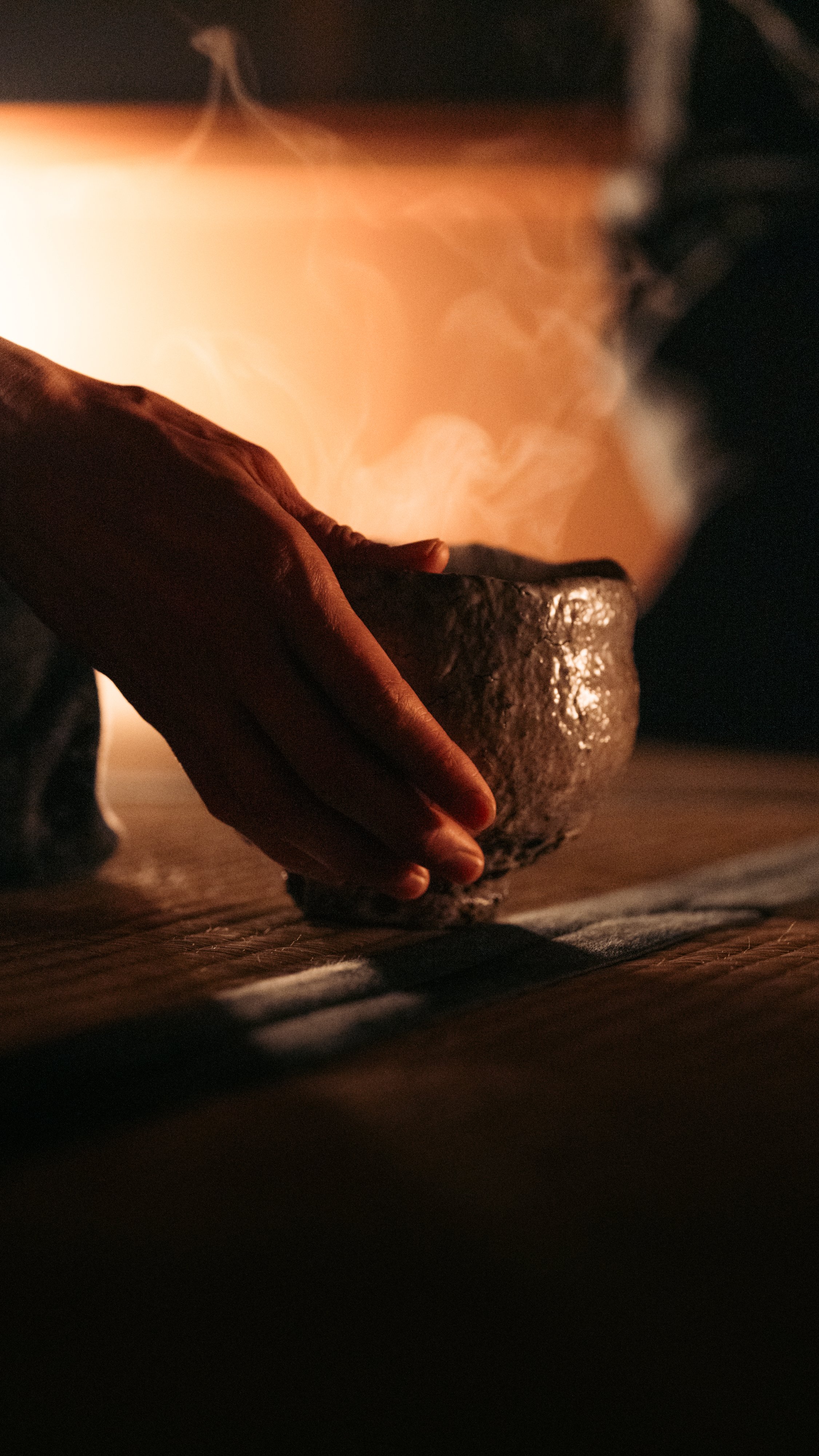 Close-up of a person's hand holding a roughly textured rock over a flame, with smoke rising, in low lighting.