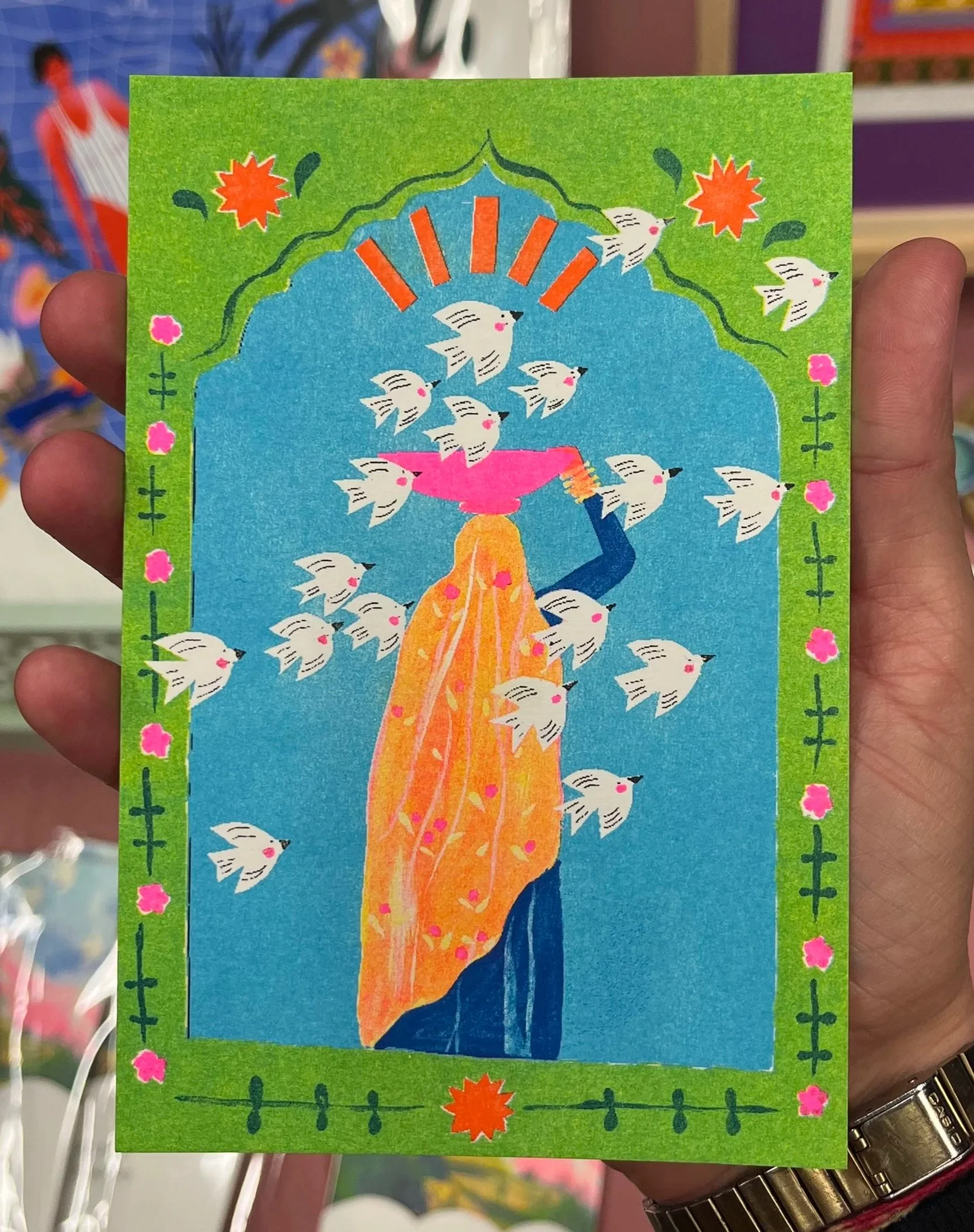 Illustration colorée représentant un arbre stylisé avec des oiseaux blancs volant autour. Le fond est bleu avec un cadre vert décoré de petits motifs floraux roses et d'oiseaux et un soleil orange en haut.