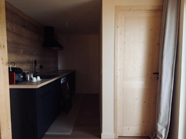 A small kitchen area with black cabinets, a wooden countertop, a sink, a black range hood, and some small kitchen appliances. There is a light-colored wooden door and beige curtains on the right side.