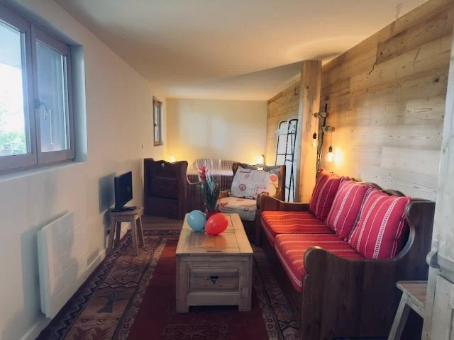 Cozy living room with wooden accent wall, red striped sofa, coffee table with potted plant and balloons, small TV, and natural light from windows.