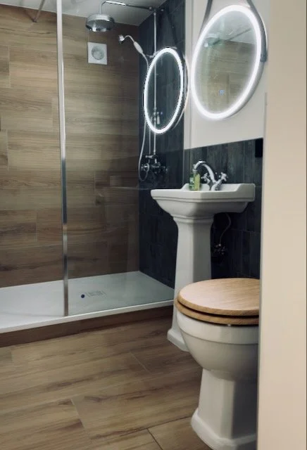 Bathroom with a walk-in shower, round backlit mirror, pedestal sink with toiletries, and a toilet with a wooden seat cover.