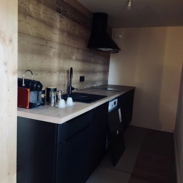 Small kitchen with black cabinets, wooden countertop, and a wooden wall panel. Contains a coffee machine, glasses, cups, a sink, and a stovetop. Black range hood above the stove.