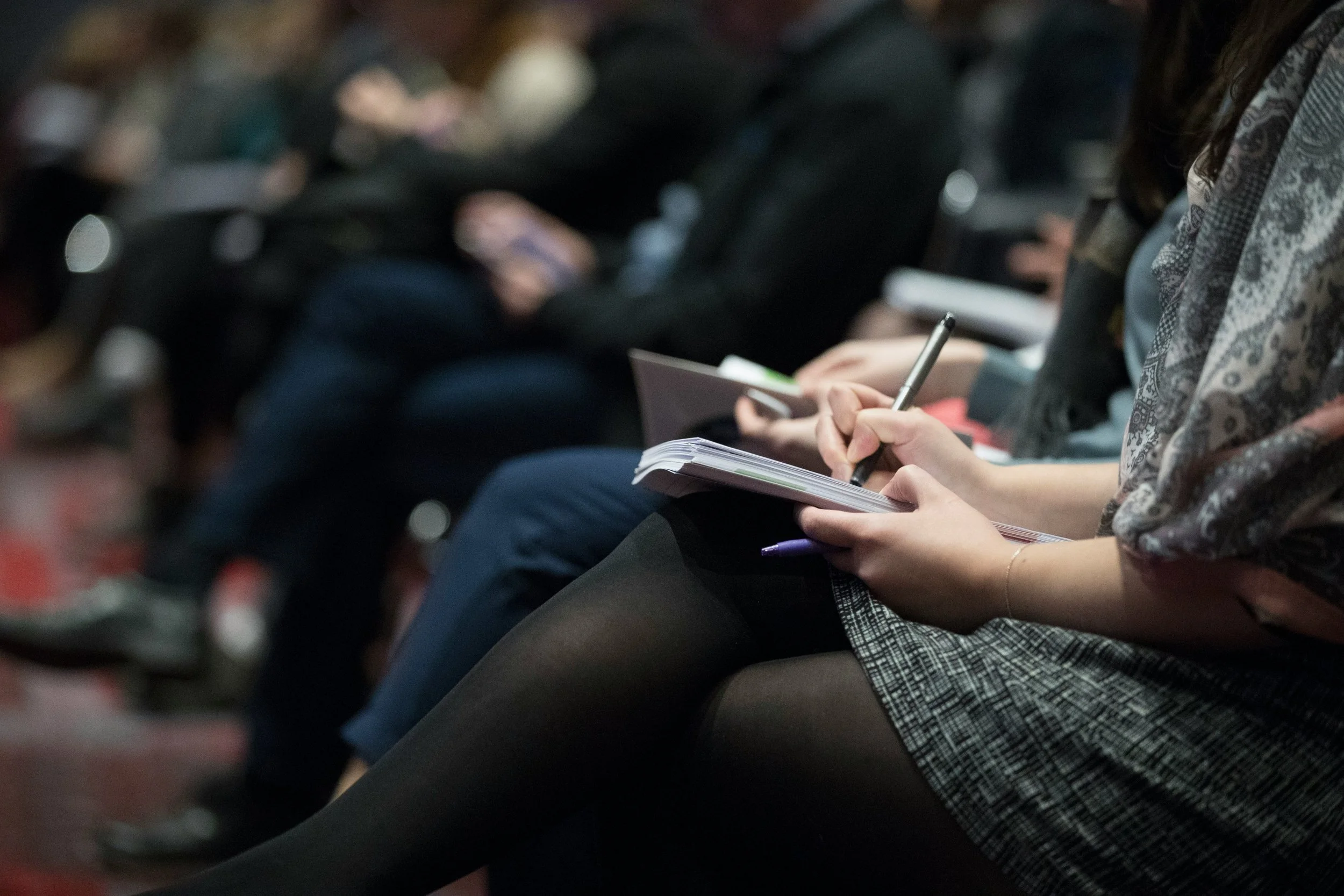 People sitting in a row at an event, taking notes in notebooks and using pens.