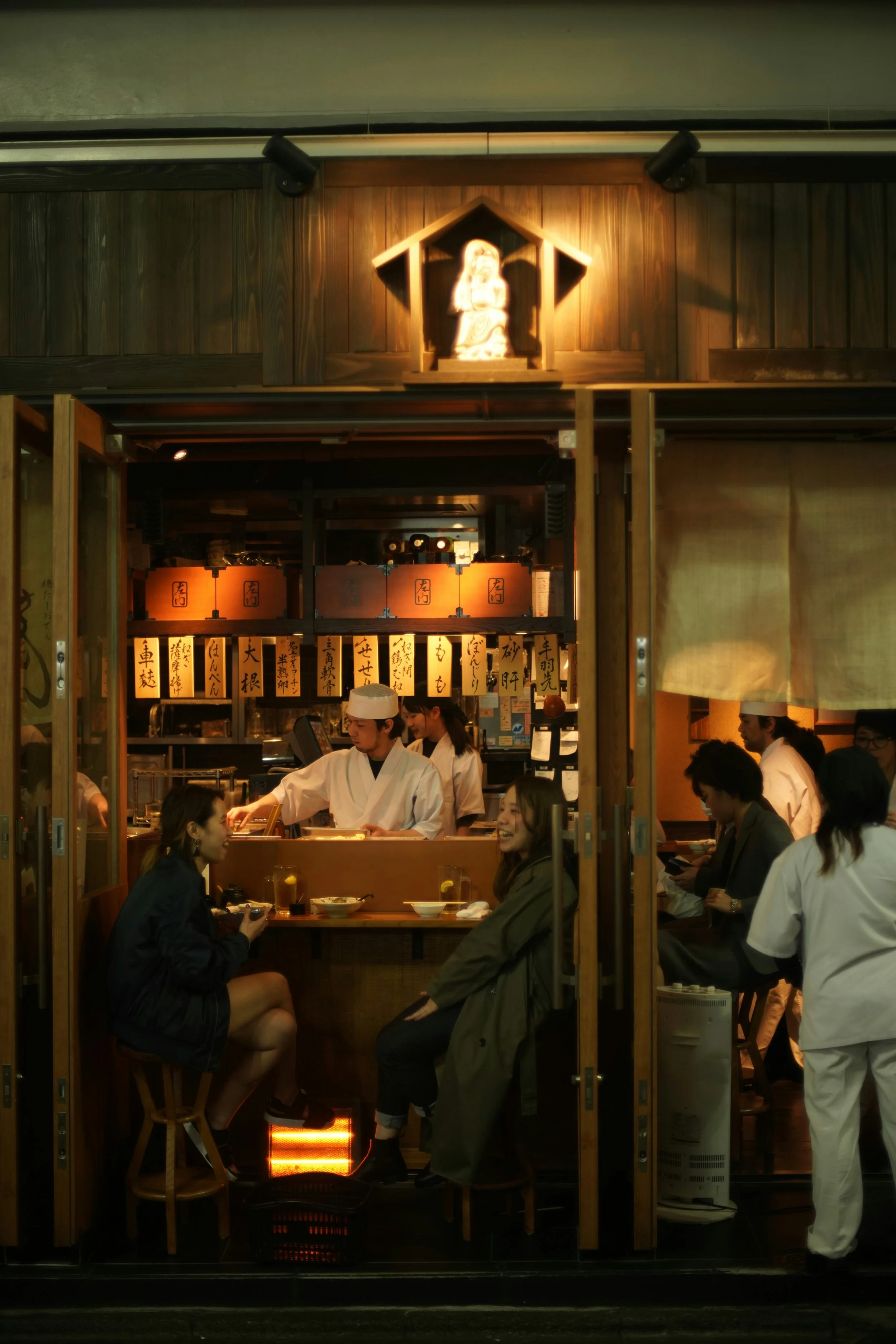 Inside a Japanese restaurant with a small counter, two women sitting and chatting at the counter, chefs preparing food behind them, and several people seated inside, all surrounded by wooden decor and traditional Japanese signs.