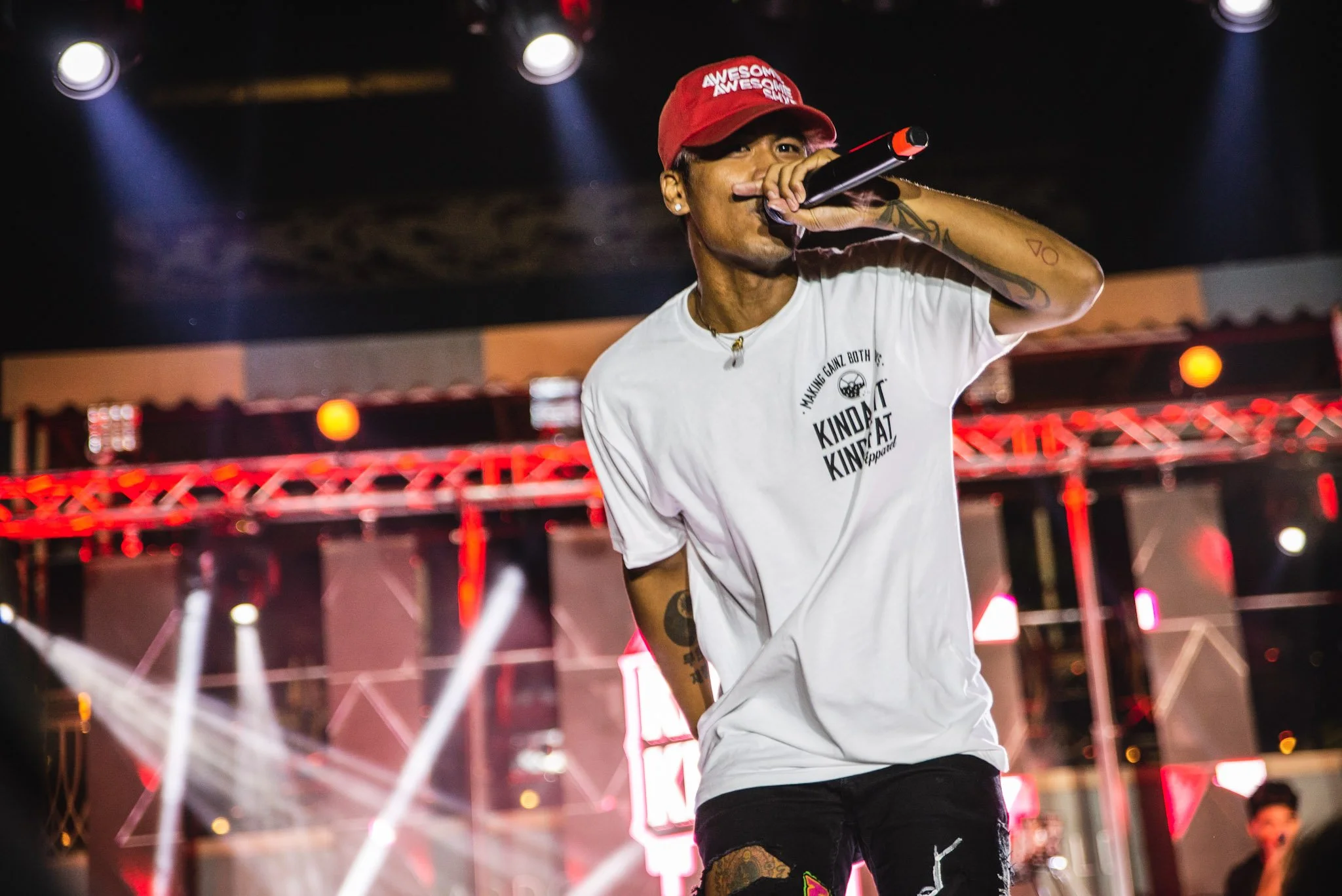 A male performer singing into a microphone on stage under bright spotlights, wearing a red cap and casual white T-shirt at a concert or event.