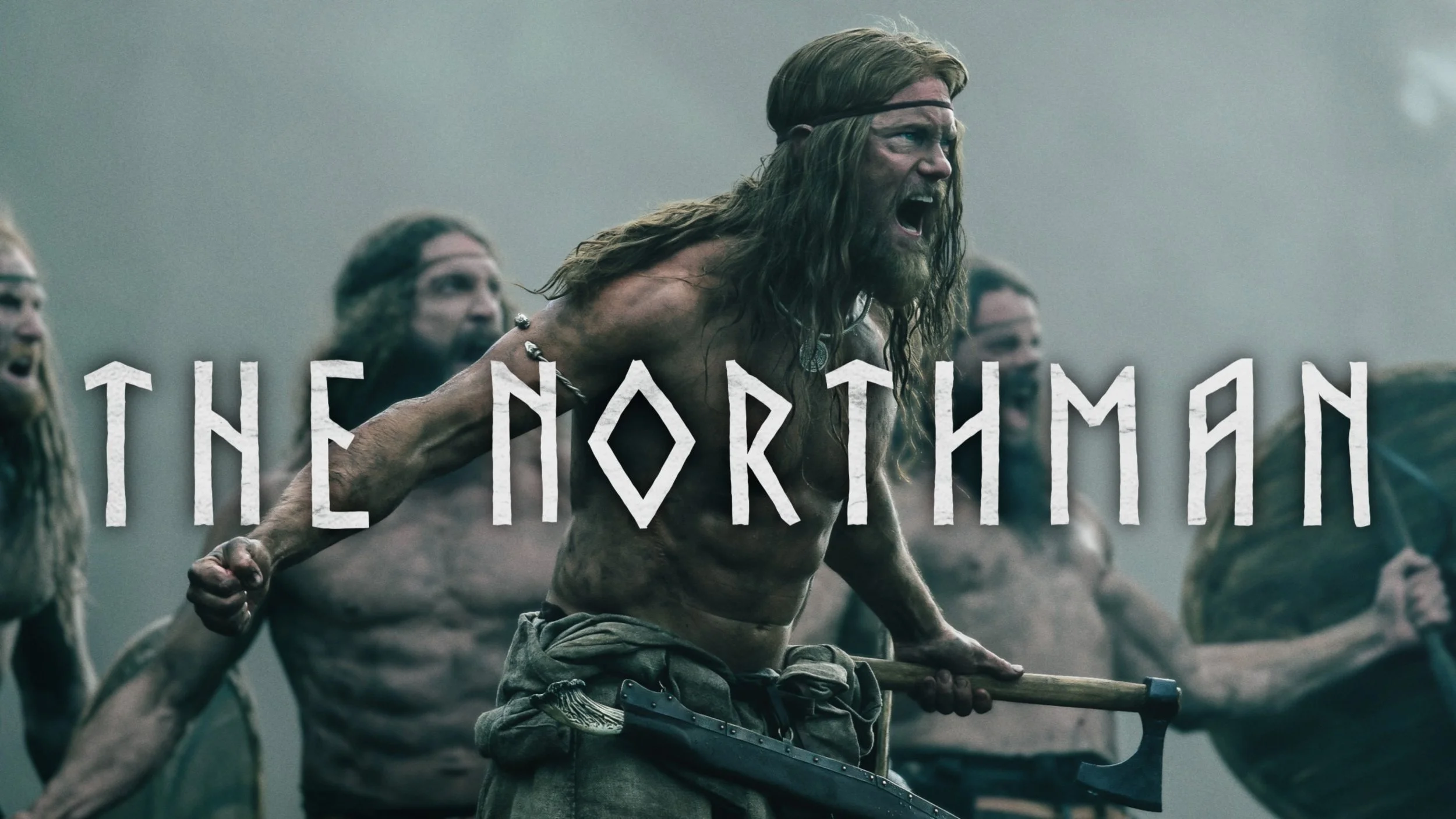 A shirtless man with long hair and a beard, wearing a headband, appears to be leading a group of similarly dressed men, all with long hair and beards, who are running or moving forward together on a foggy field. The text 'THE NORTHMAN' is overlaid on