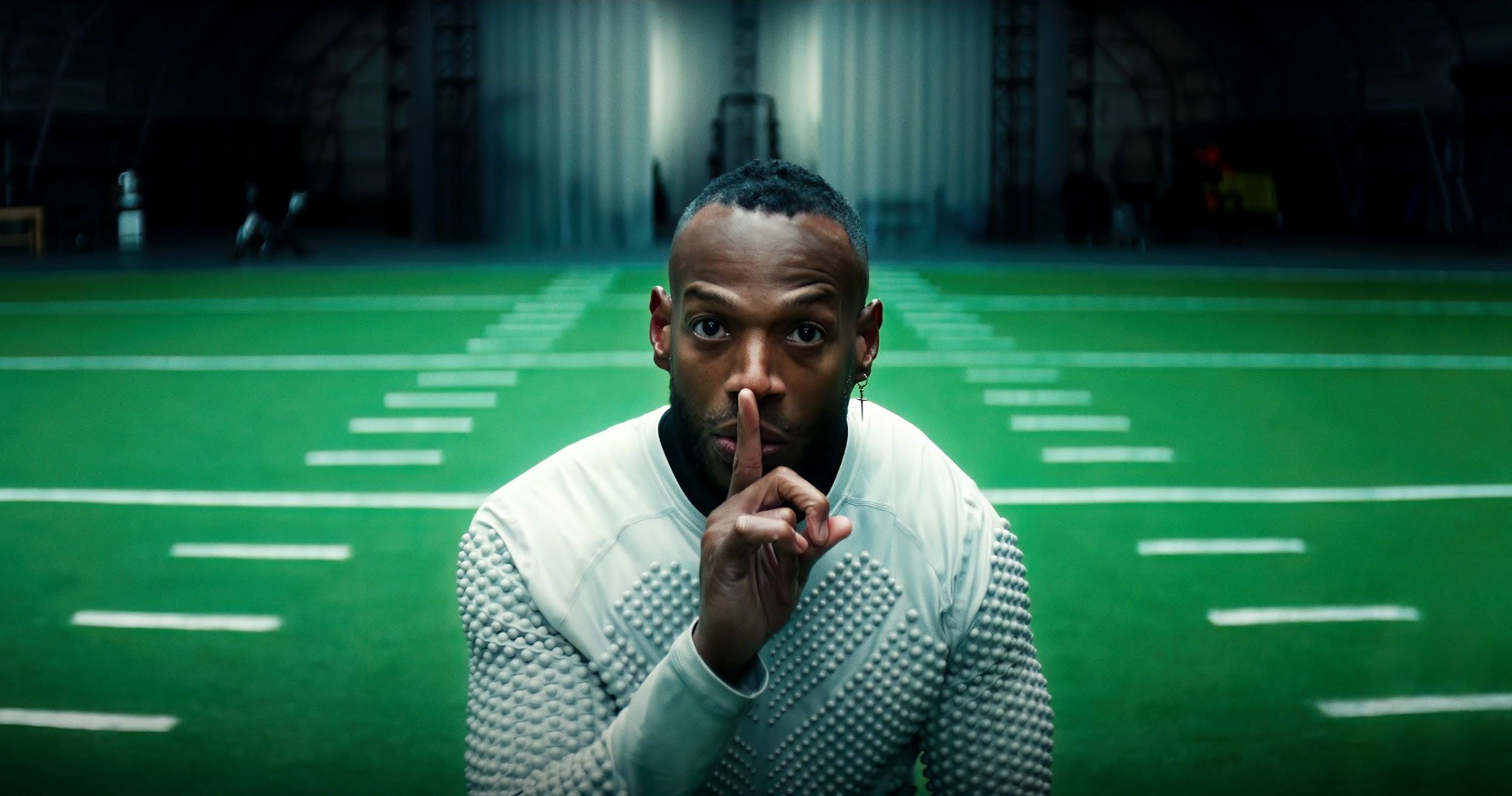 Man kneeling on a football field with his index finger over lips, in an indoor sports facility.
