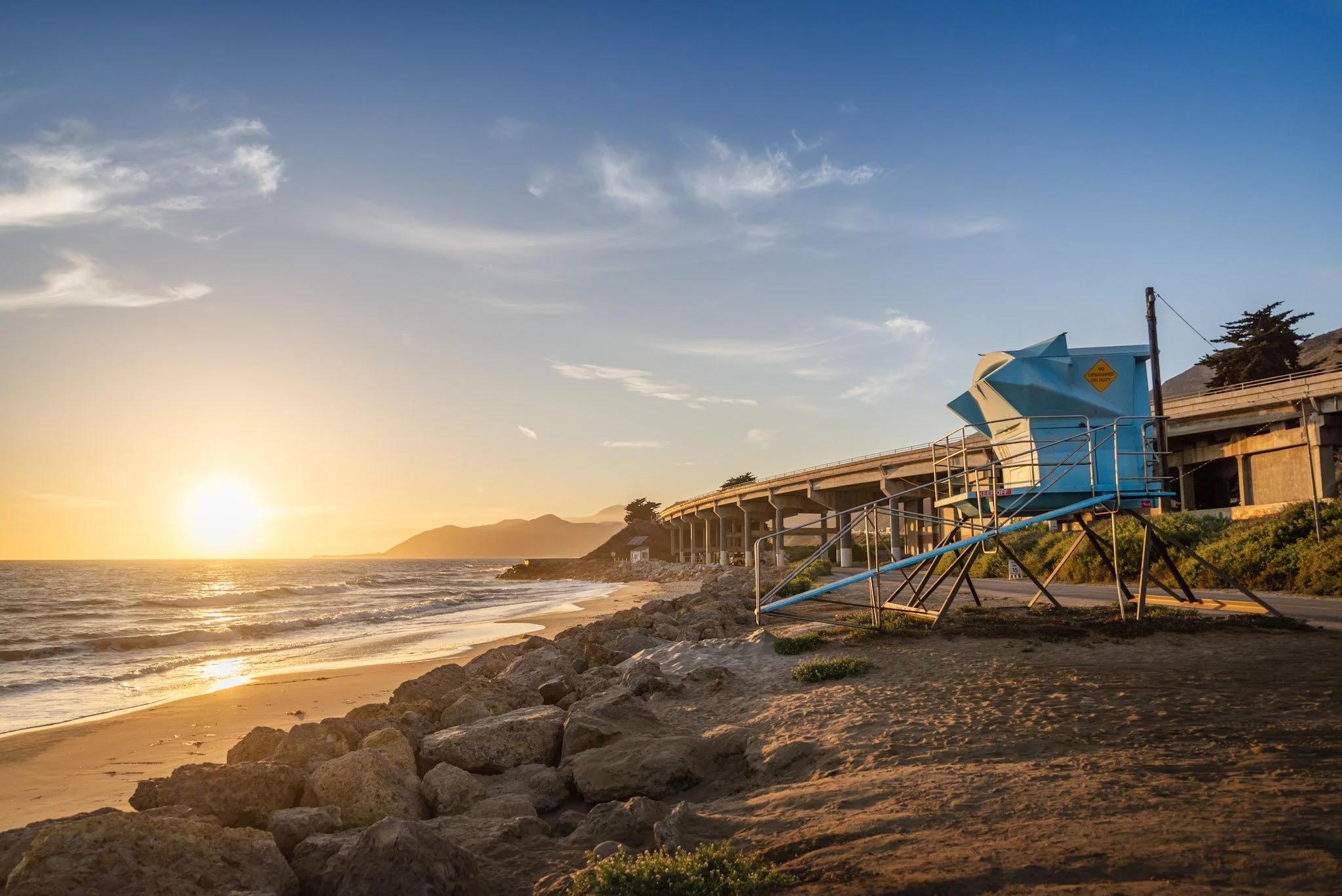 Sunset over a beach with rocky shoreline and lifeguard tower.