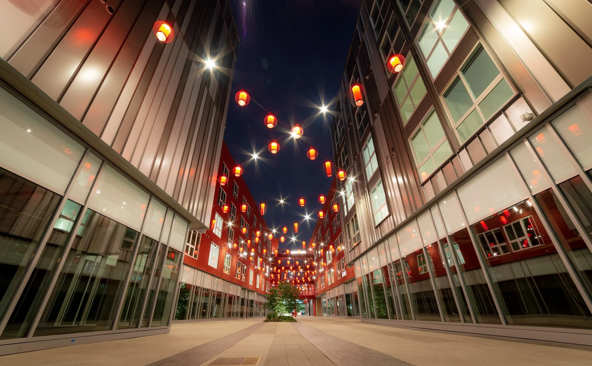 Night view of a modern urban street with glass buildings on both sides and red lanterns hanging overhead.