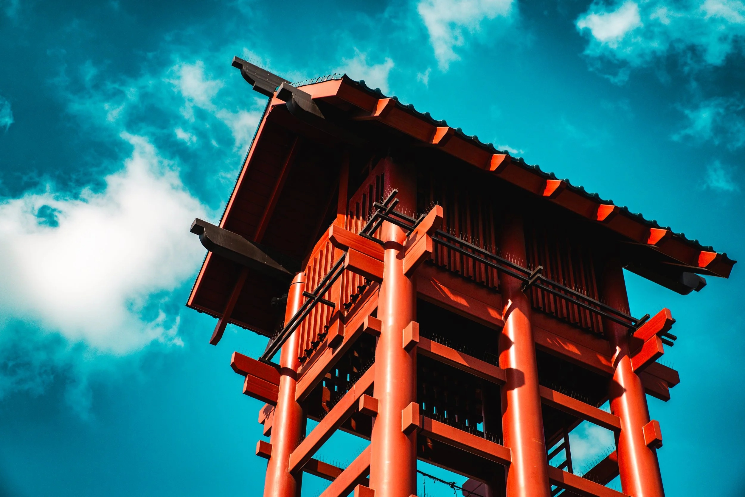 A traditional red Japanese pagoda with intricate wooden architecture and curved roof, set against a bright blue sky with scattered clouds.