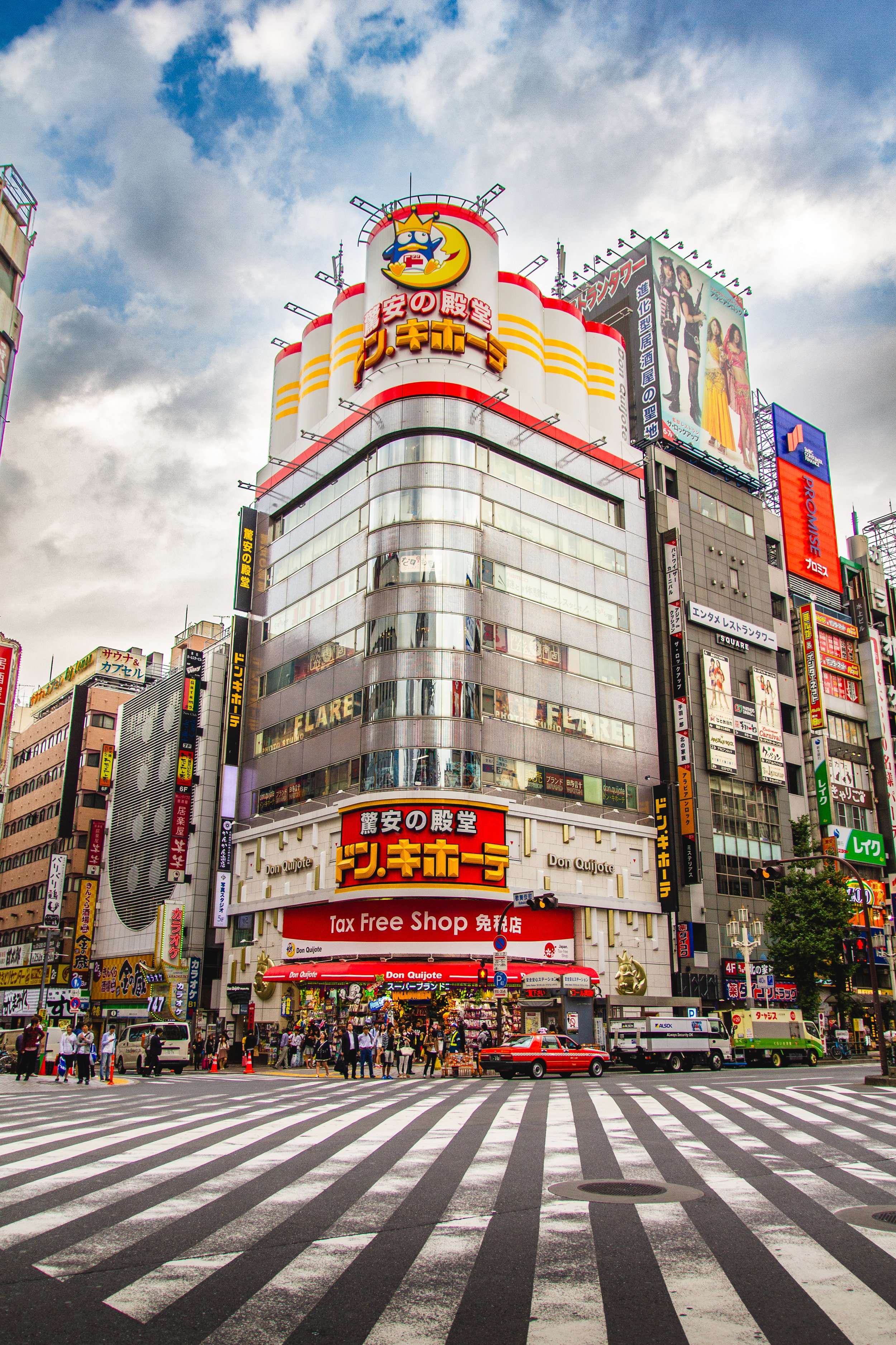 A busy city intersection with a tall building featuring various signs in Japanese, including a prominent Don Quijote store. The scene shows pedestrians crossing the street, cars, and trucks. The sky is cloudy with some patches of blue.