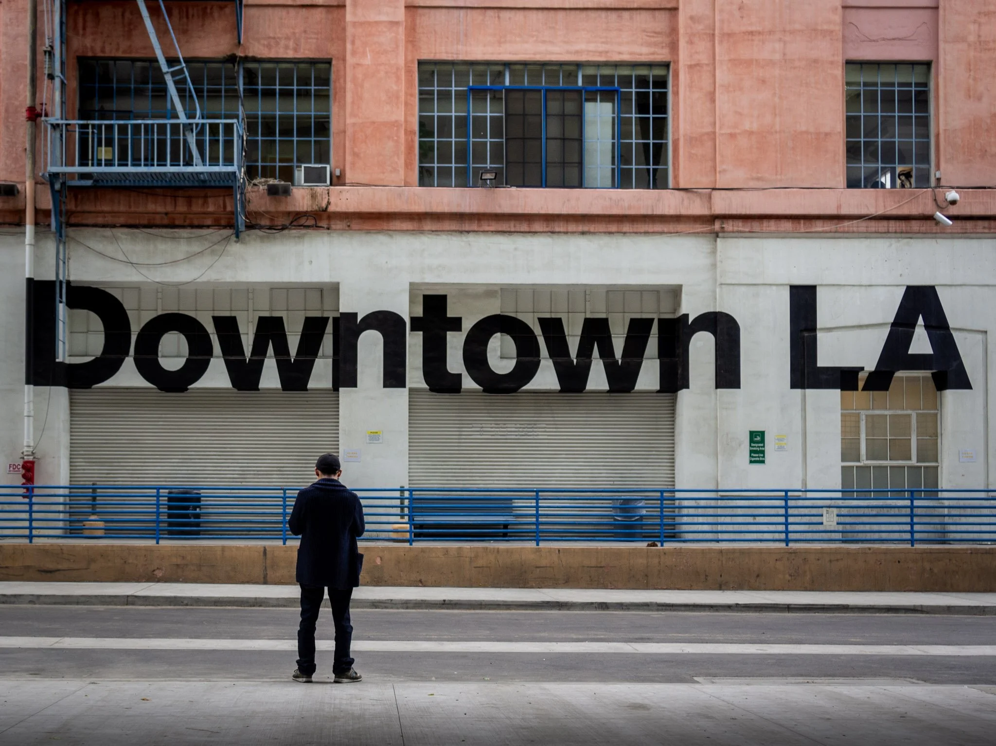 A man standing on the sidewalk, facing away, looking at a large building with a sign that partially reads 'Downtown LA' and a row of blue barrels in front.