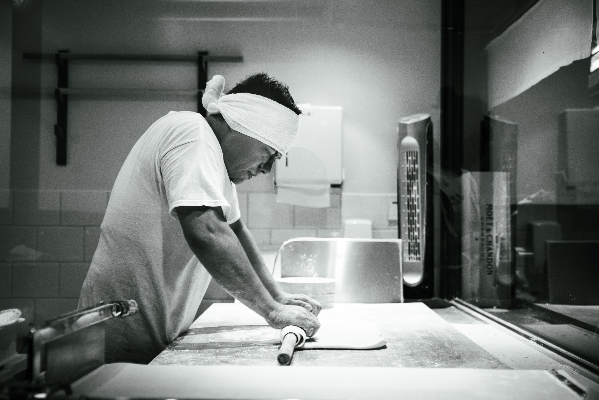 A person in a white T-shirt and a headband is rolling out dough with a rolling pin in a kitchen.
