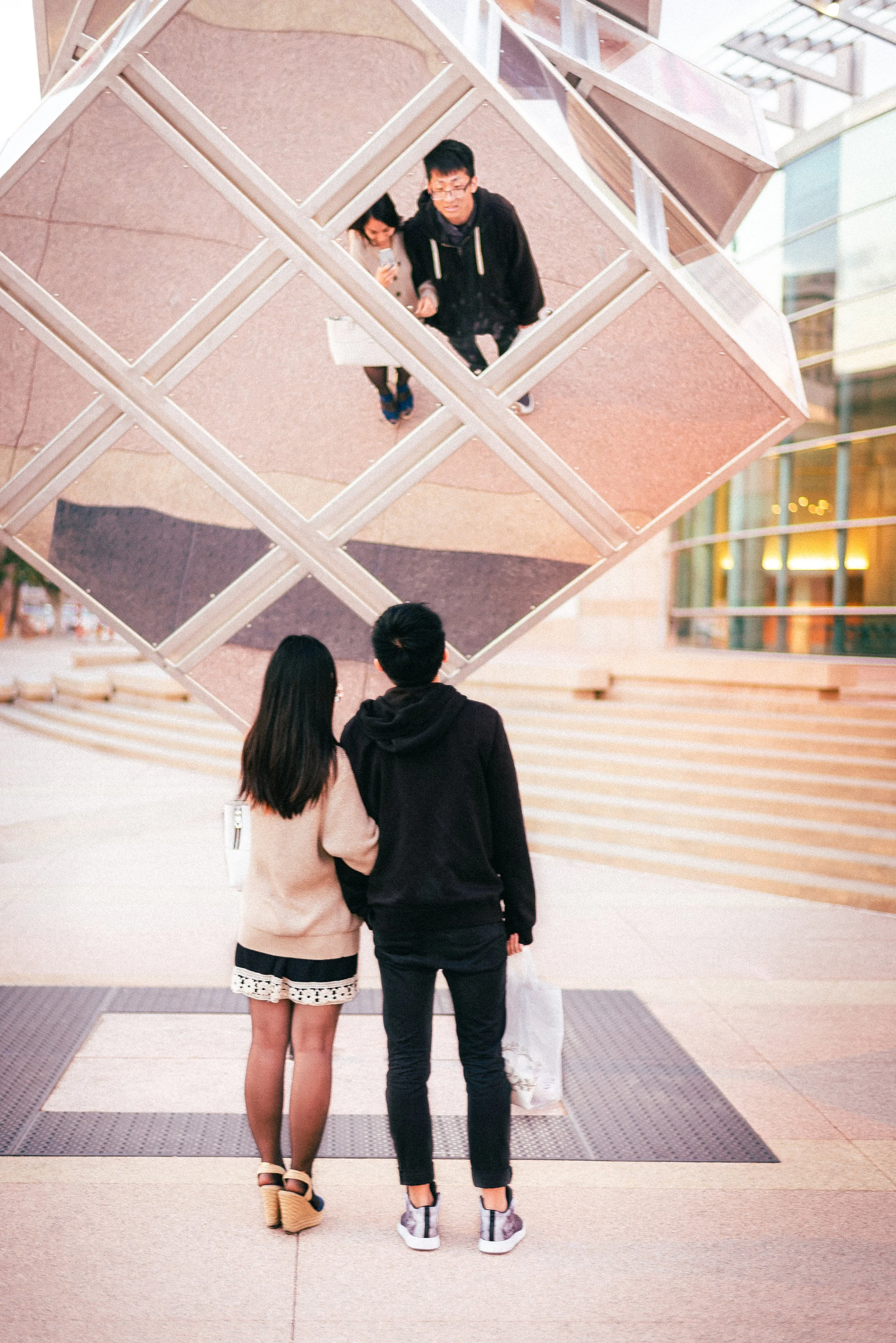 Two women and one man standing outdoors, viewed in a mirror that is part of an architectural structure with diamond-shaped reflections. They are looking at their reflections and appear to be taking photos or selfies.