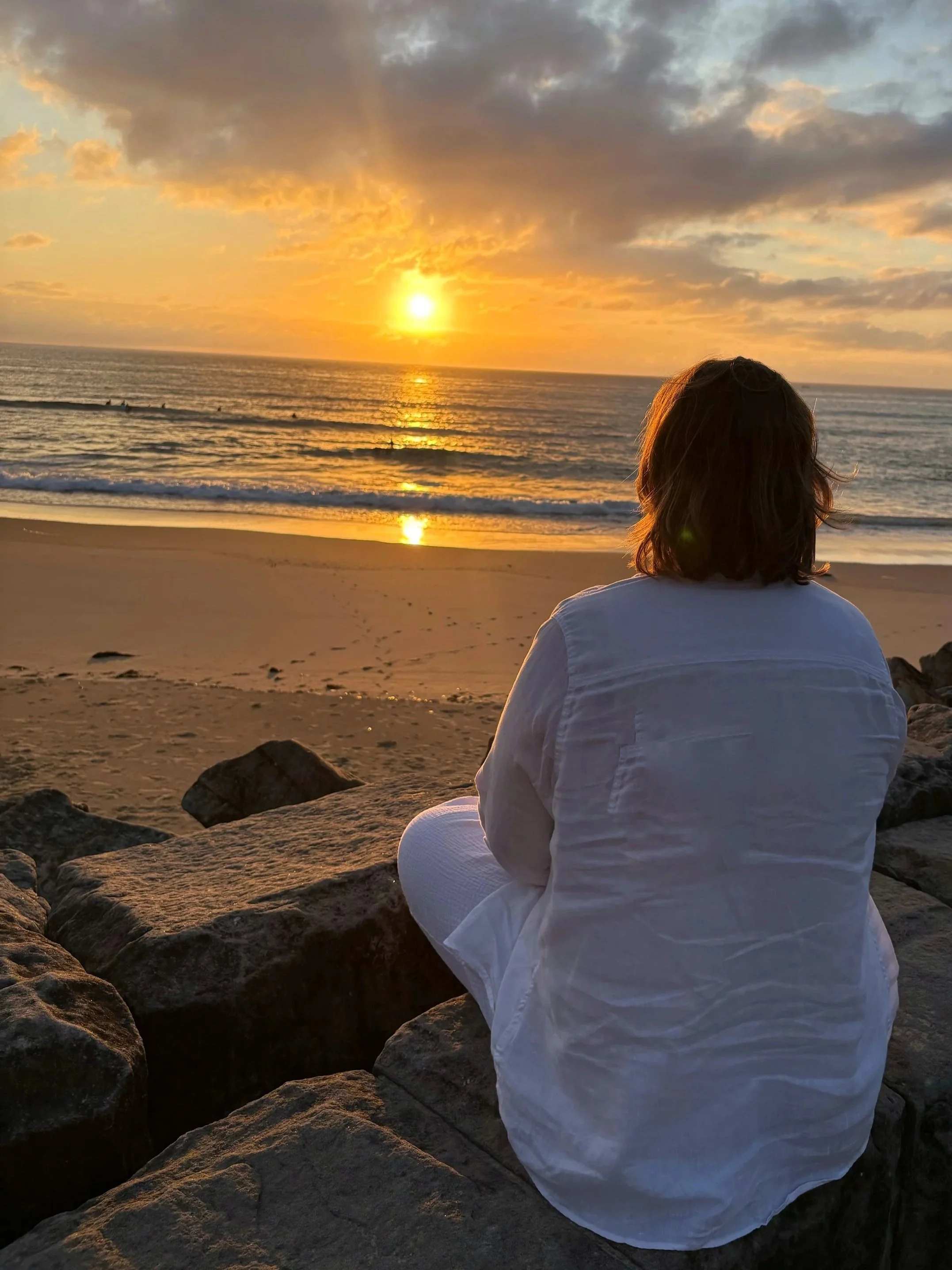 Person reflecting on life and sitting on rocks by the beach, watching the sunset and connecting with self