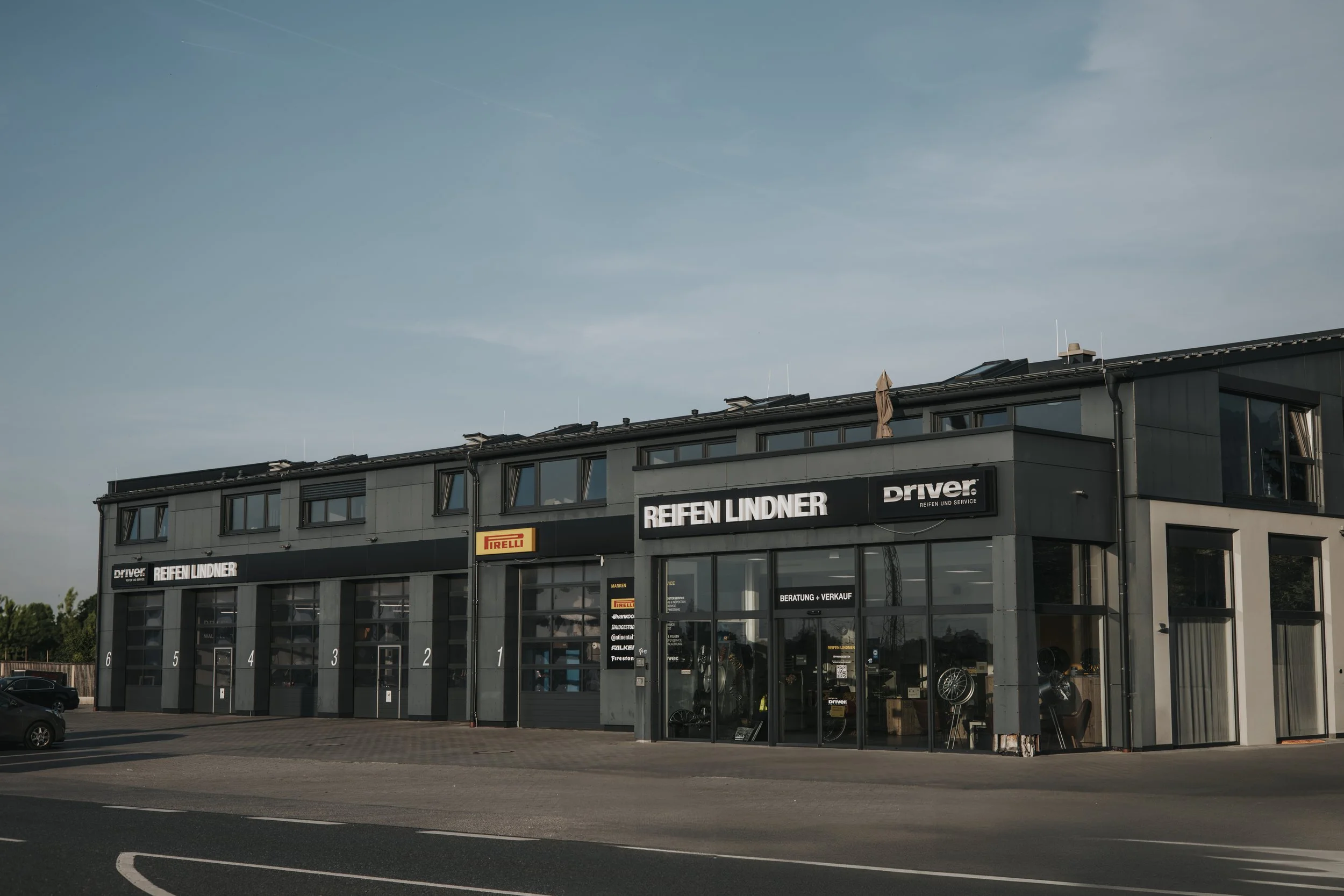 Außenansicht eines modernen Reifenfachgeschäfts mit mehrer Shops, große Glasscheiben und Firmenlogos von Reifenmarken wie Pirelli und Firestone.
