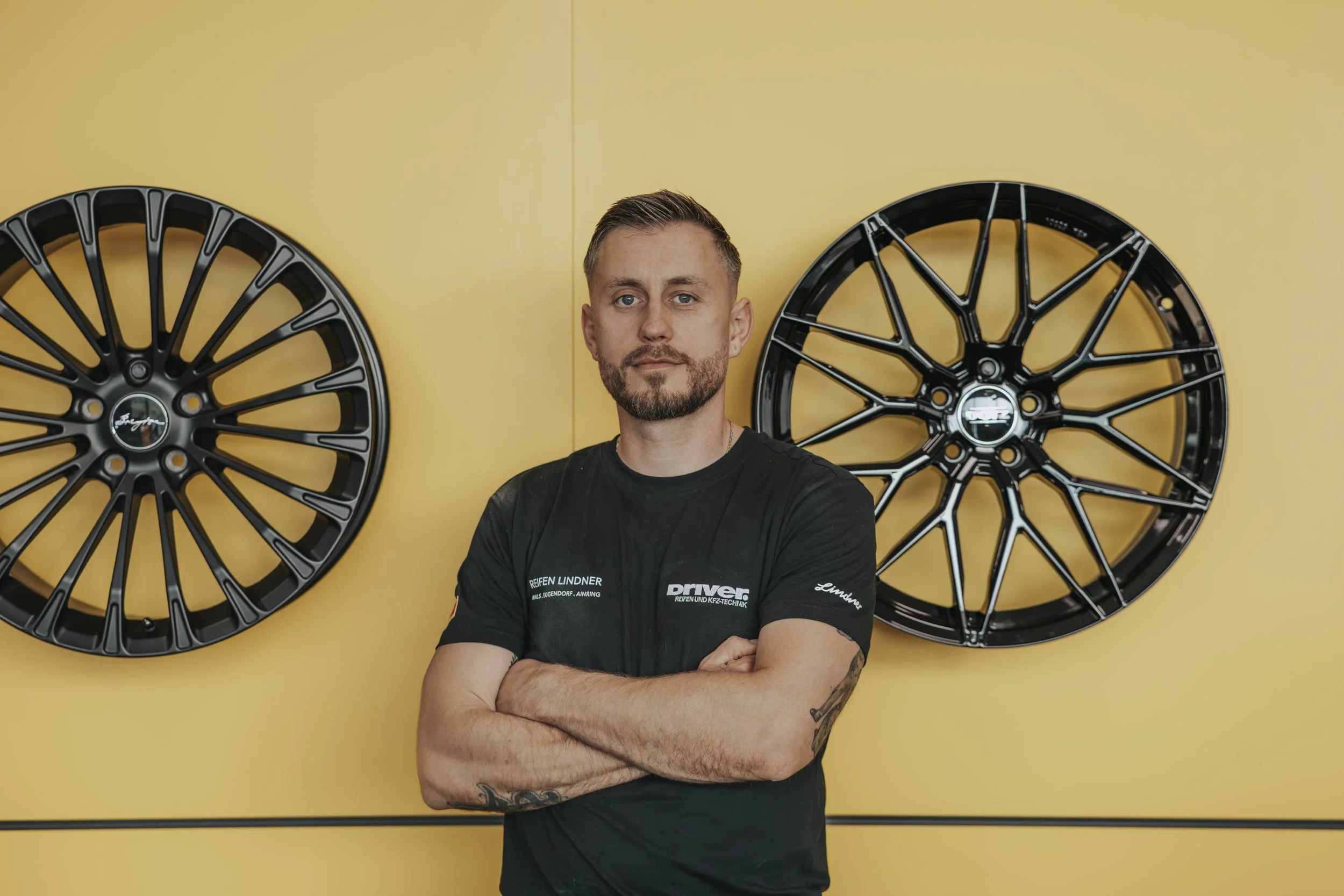 Mann steht mit verschränkten Armen vor gelbem Hintergrund mit zwei schwarzen Felgen an der Wand.