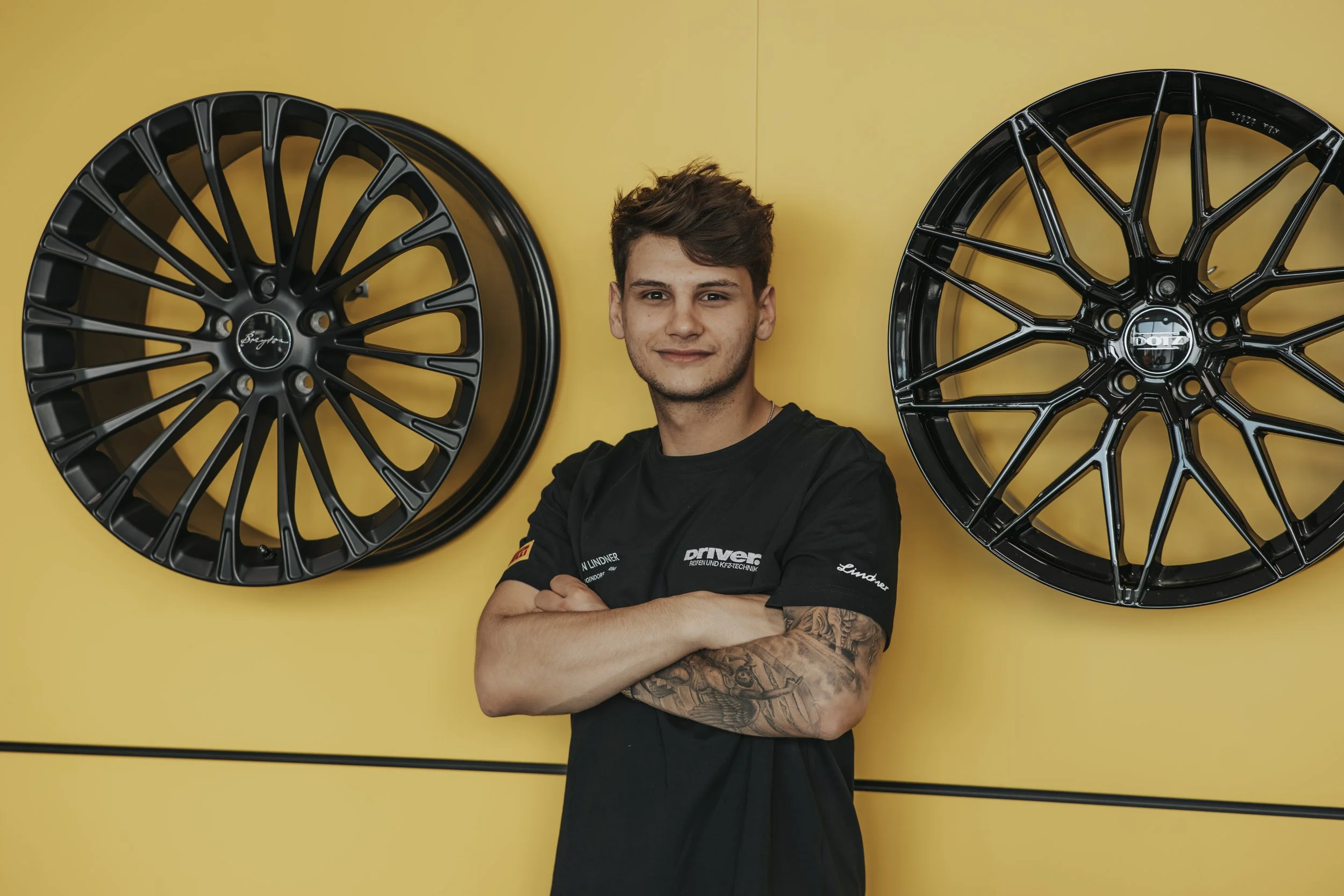 Junger Mann steht vor einer gelben Wand mit zwei schwarzen Autoreifen an der Wand, Arme verschränkt, lächelnd.
