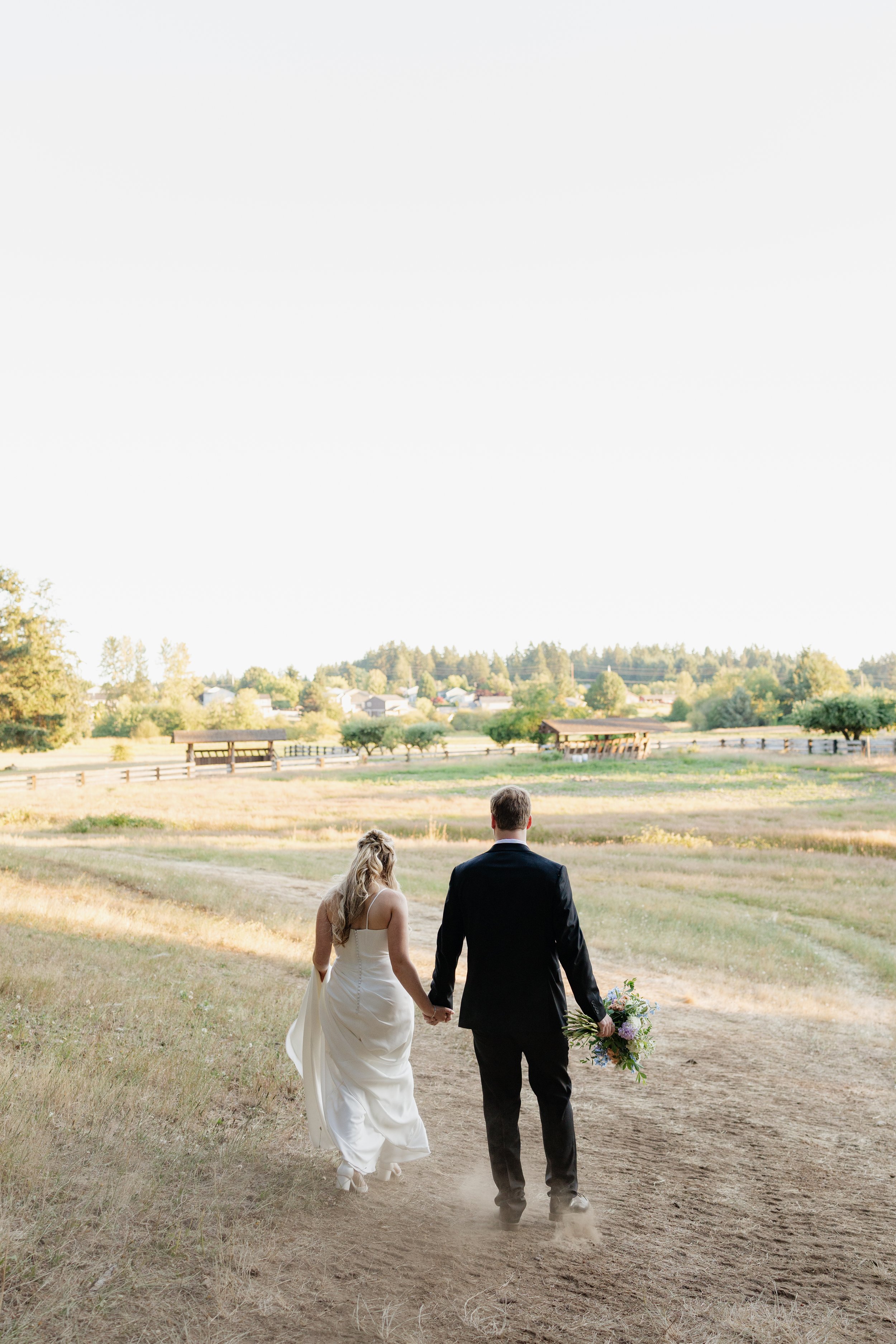 A couple, one woman in a white wedding dress and one man in a black suit, walking hand in hand on a dirt path in a grassy countryside setting, with trees, a fence, and houses in the background. The woman is holding a bouquet of flowers.