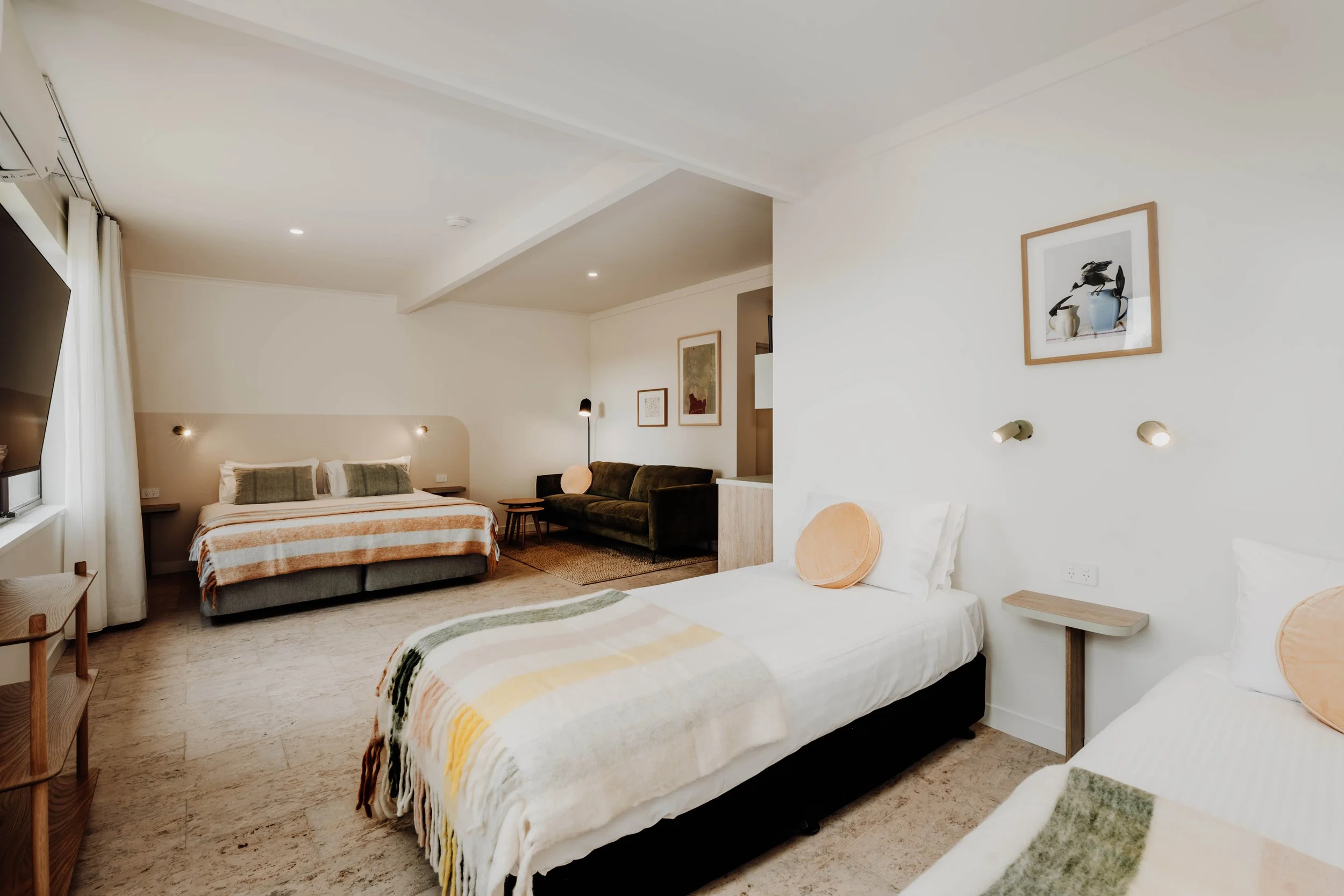 A modern hotel room with two beds, a sofa, and wall art in neutral tones, with natural lighting and minimal decor.