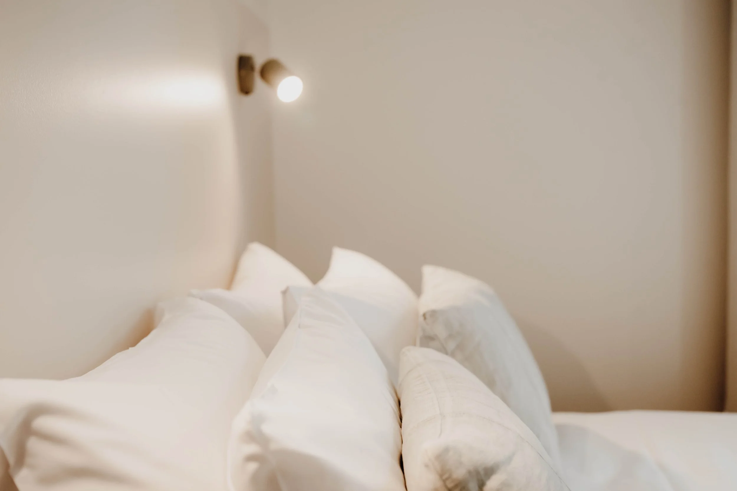 A neatly made bed with several white pillows in a bedroom with beige walls and a wall-mounted reading light.