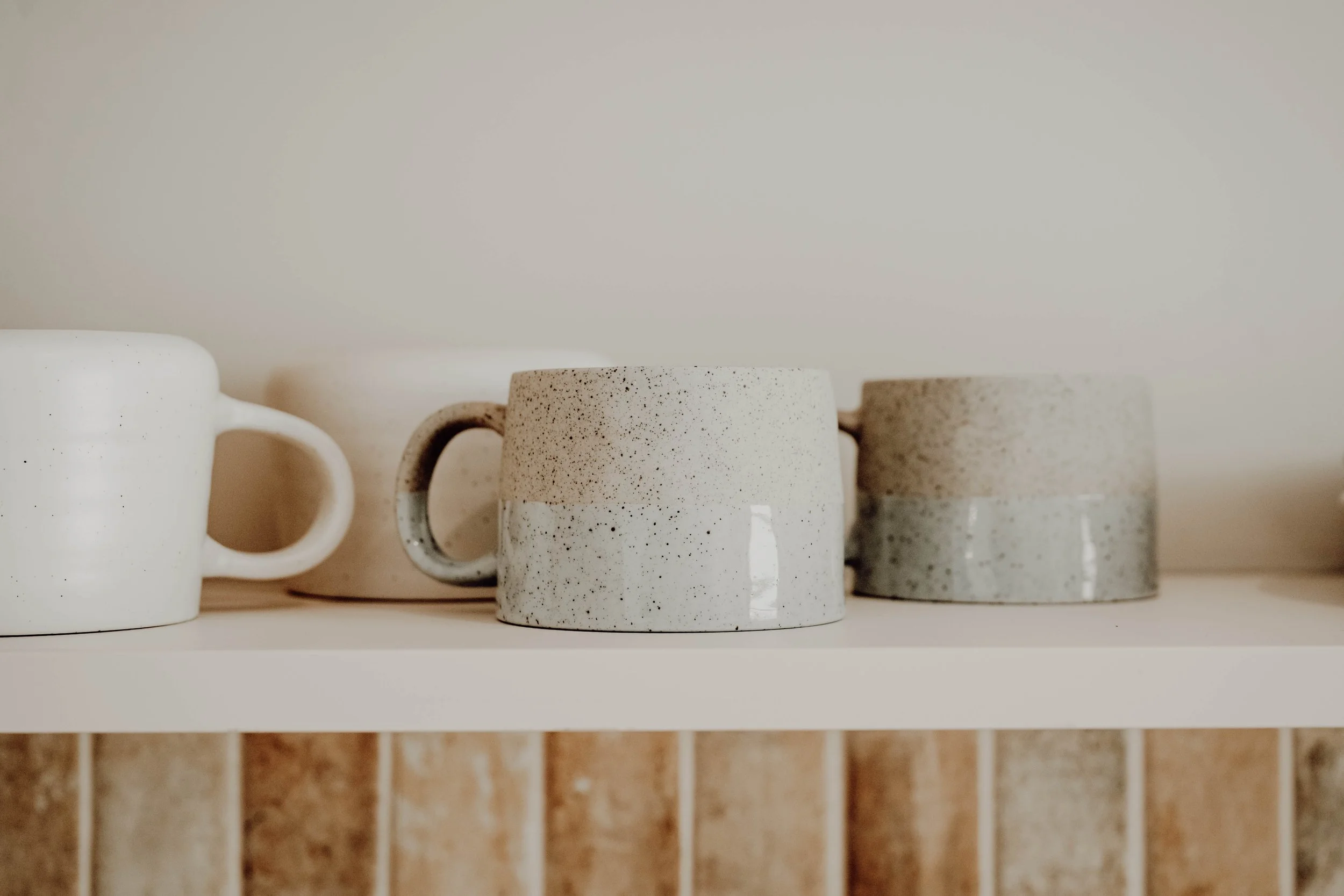 Six ceramic mugs with varying shades of beige and speckled textures are placed on a white shelf against a plain wall.