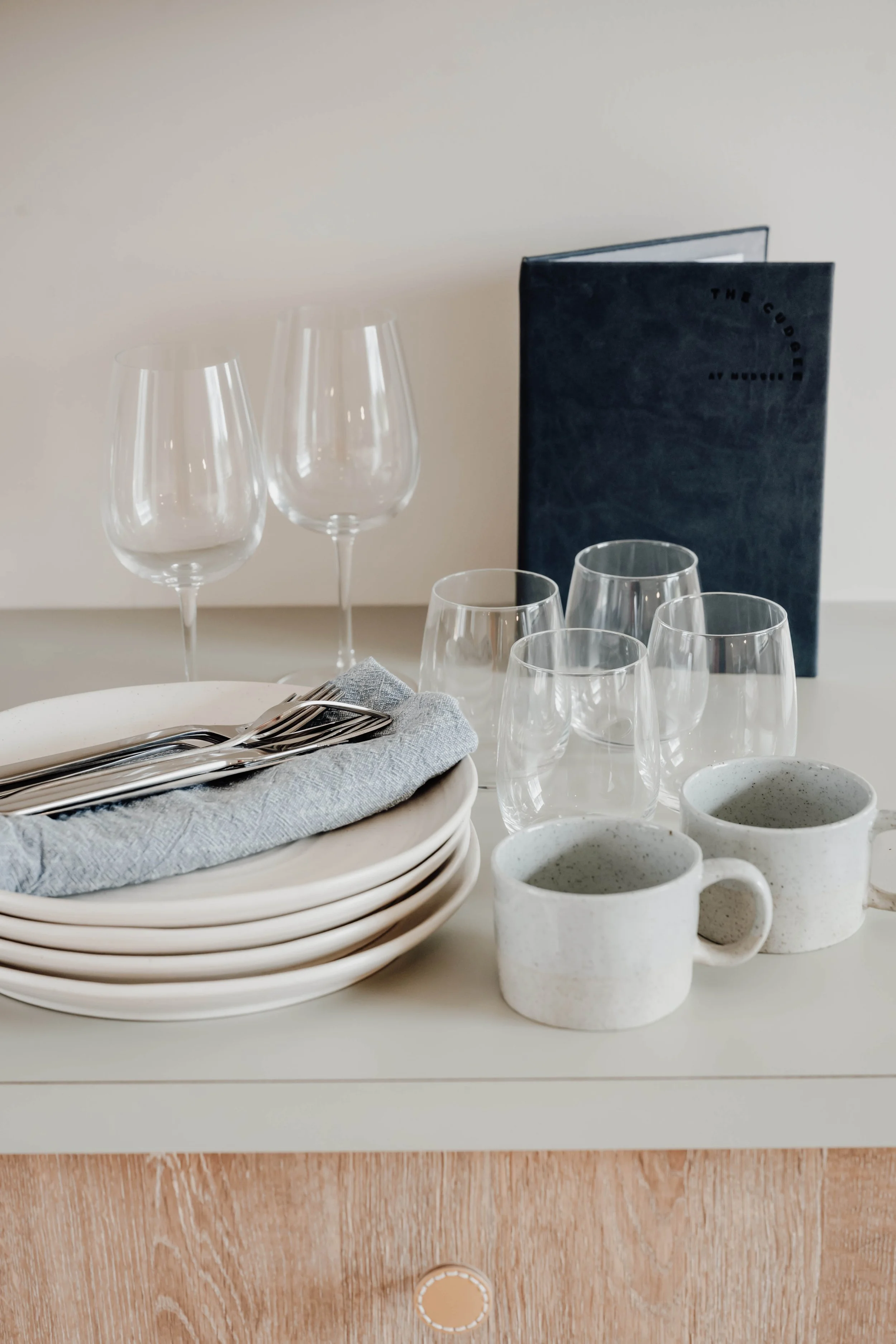 A table setting with stacked white plates, knives on a gray cloth napkin, four empty wine glasses, four plain glass tumblers, two speckled coffee mugs, and a dark blue menu or book standing upright in the background.