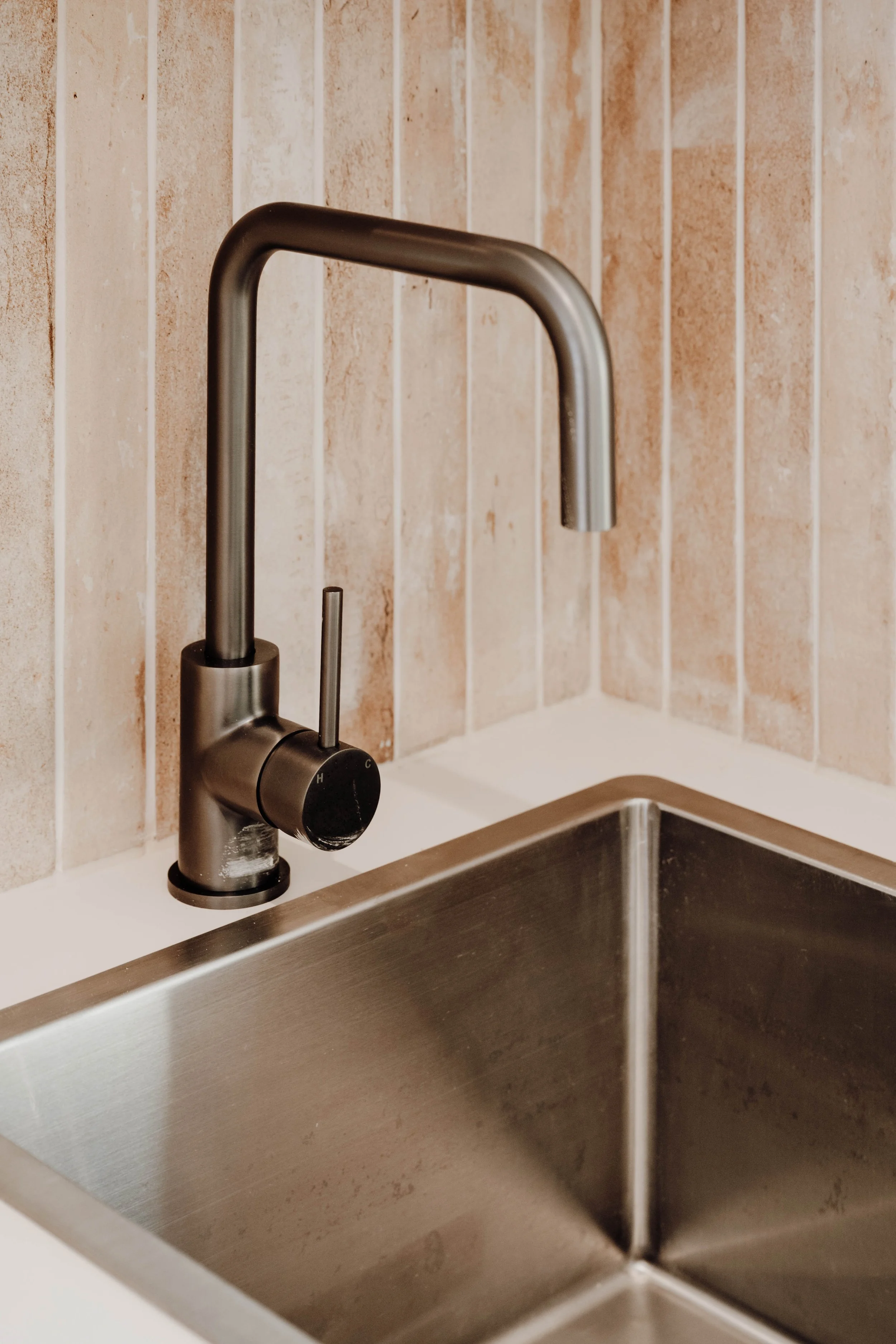 Close-up of a stainless steel kitchen sink with a modern, curved faucet and a single lever handle, mounted on a tiled backsplash with beige and light brown tiles.