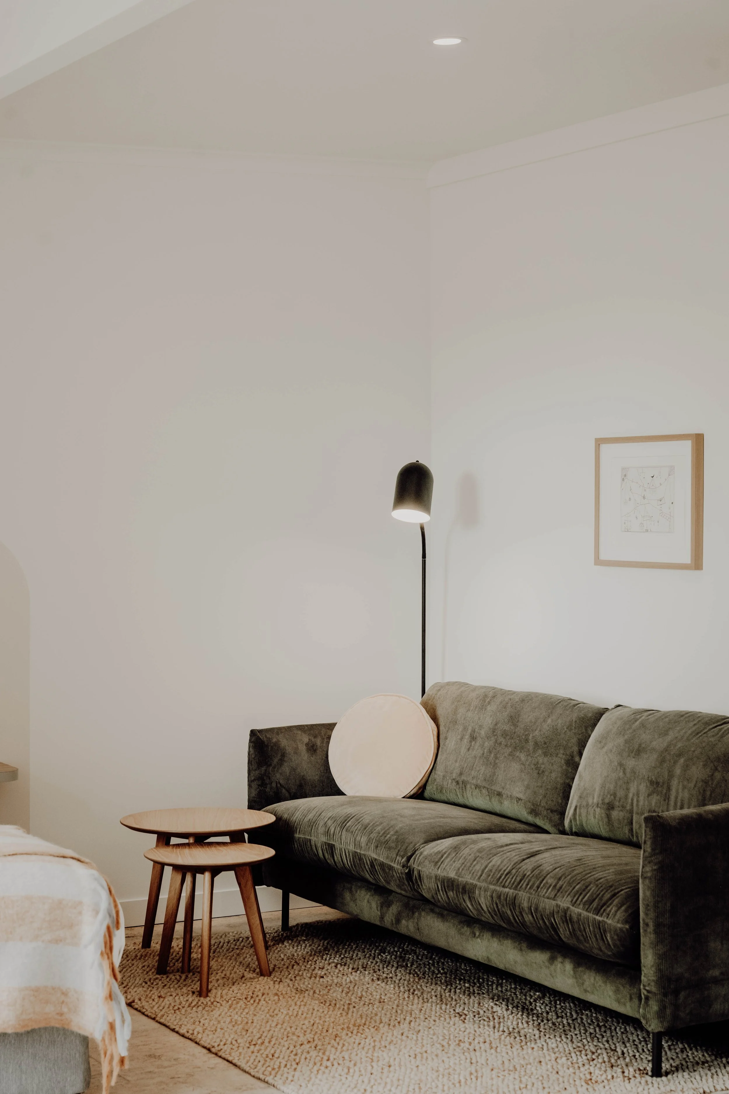 A living room with a dark green velvet sofa, a round pink cushion, two small wooden nesting tables, a beige textured rug, a tall black floor lamp, a framed artwork on the wall, and part of a bed with striped bedding visible in the corner.