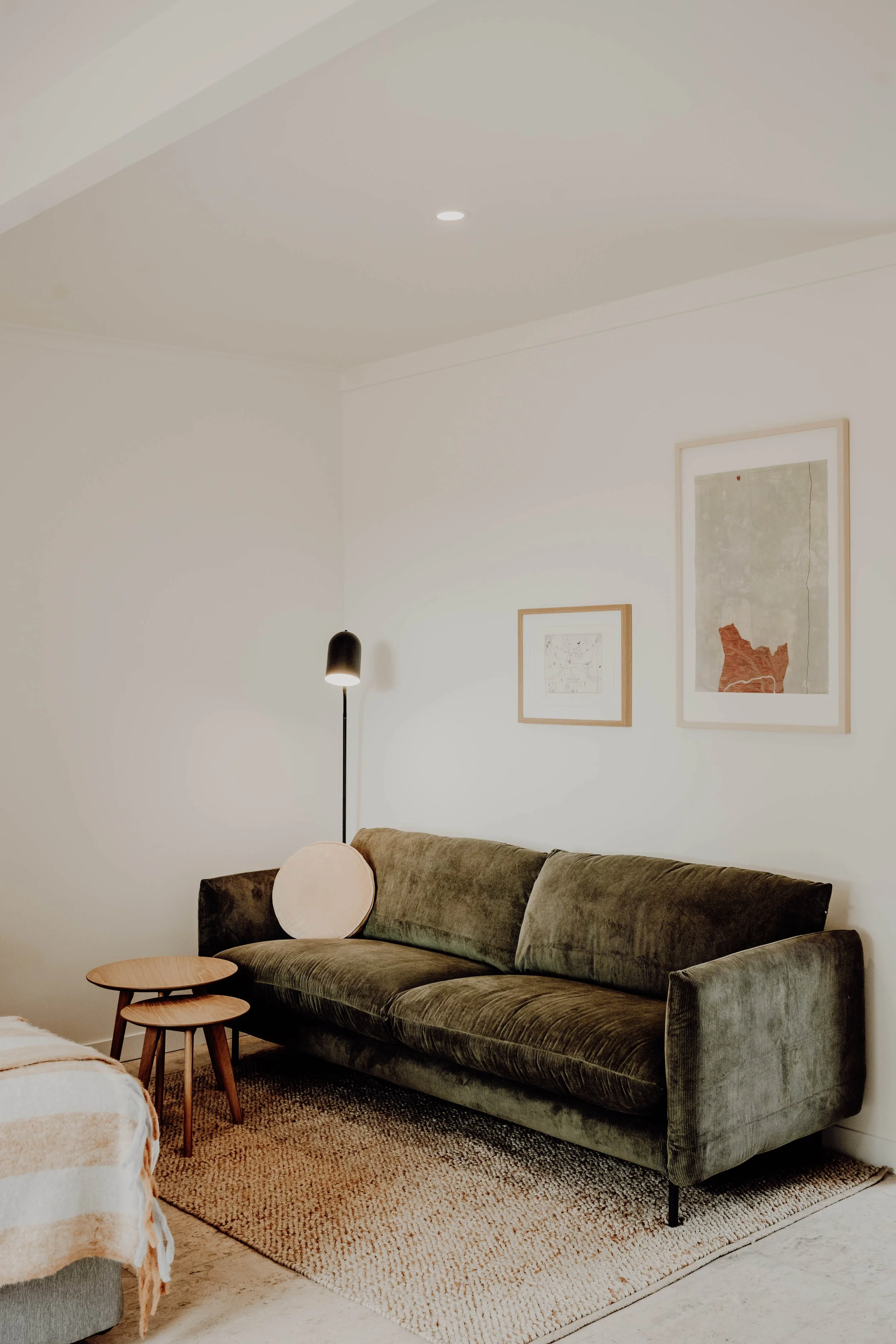 Living room with green velvet sofa, small nested wooden tables, framed artwork on white wall, black floor lamp, beige area rug, and a part of a bed in the foreground.