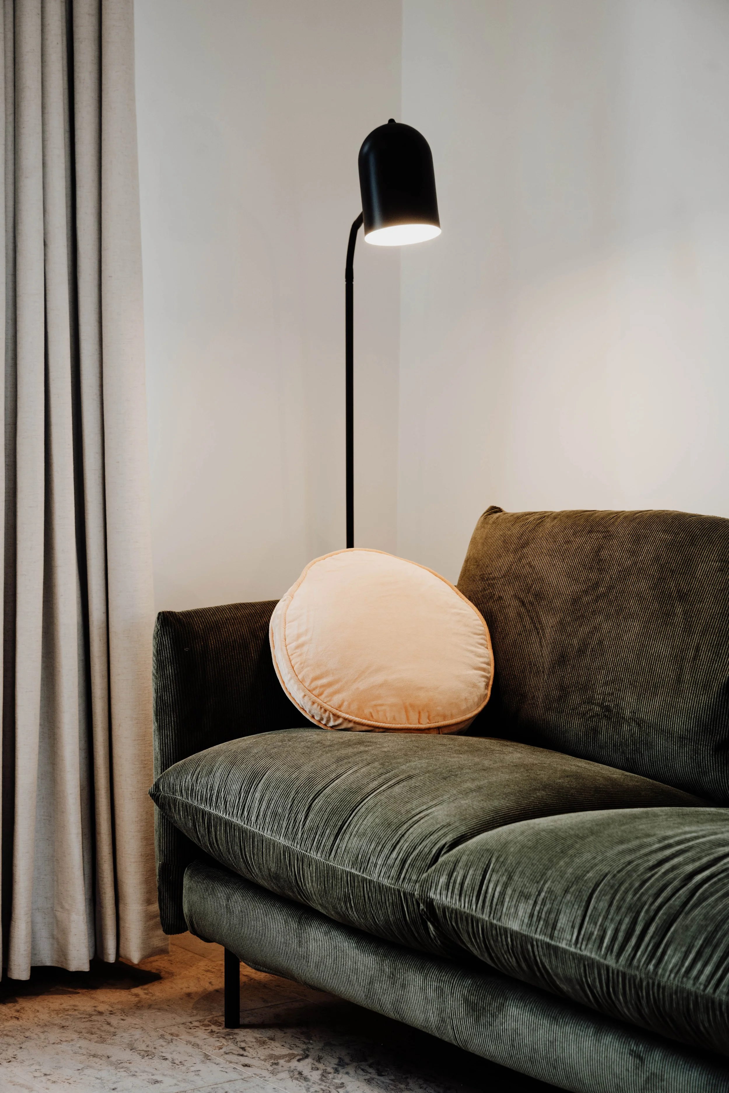 A dark green velvety sofa with a peach-colored round pillow and a black floor lamp with an upward-facing shade in a cozy living room corner.