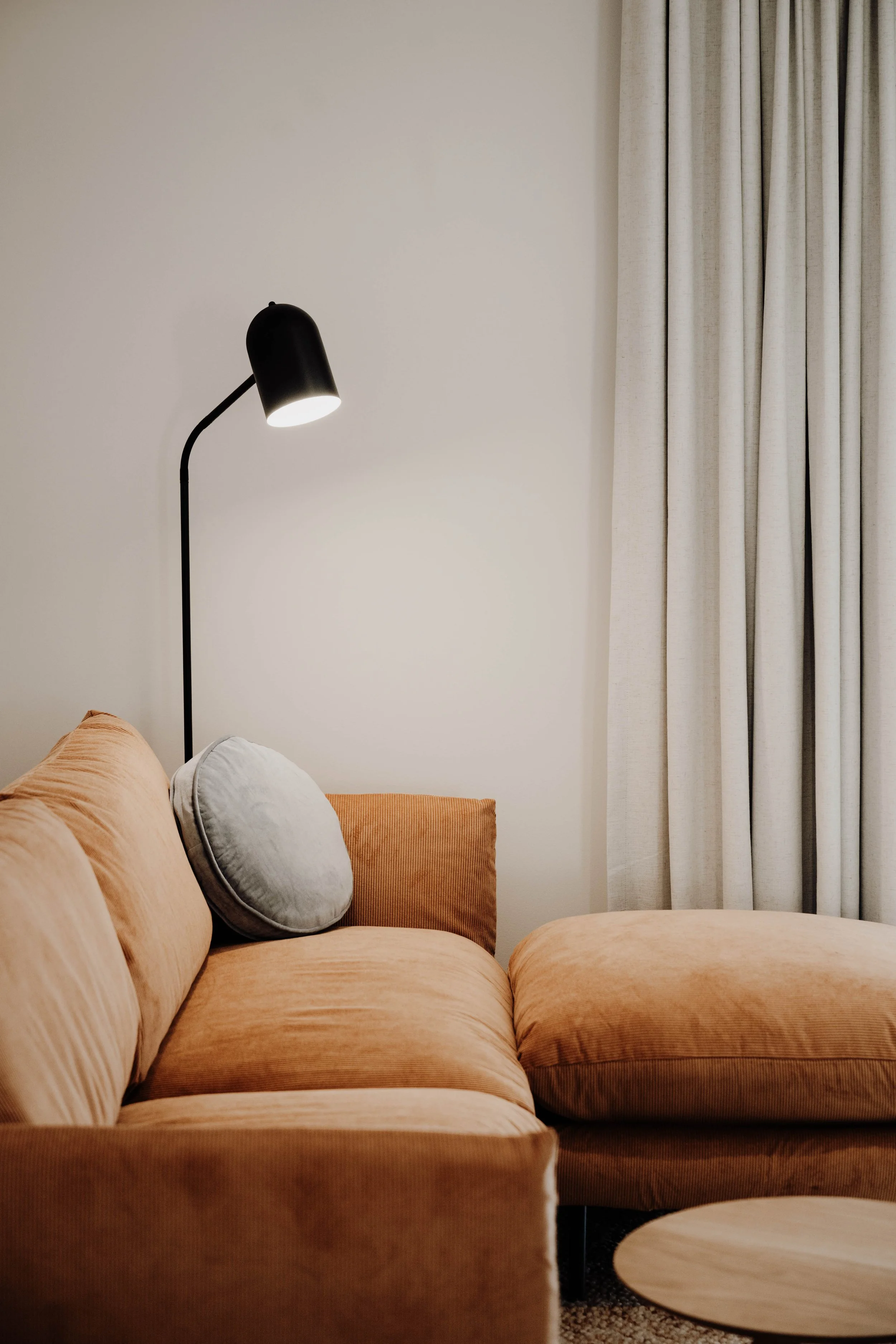 A living room corner with an orange sectional sofa, a round gray pillow, a black floor lamp, a white curtain, and a light-colored wall.
