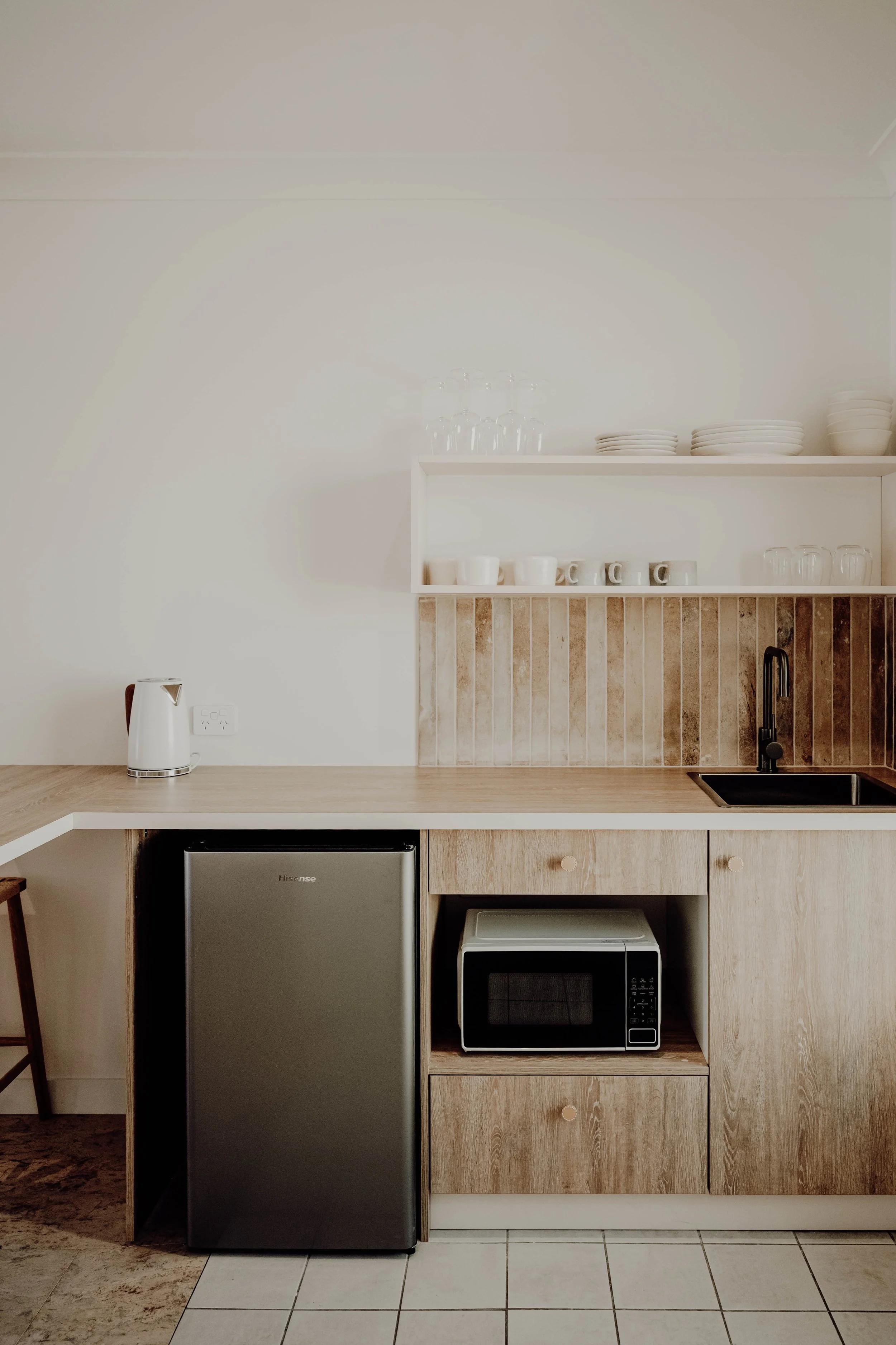 A minimalist kitchen with light wooden cabinets, a small refrigerator, a microwave, a black sink with a black faucet, and white shelves holding plates, cups, and glassware.