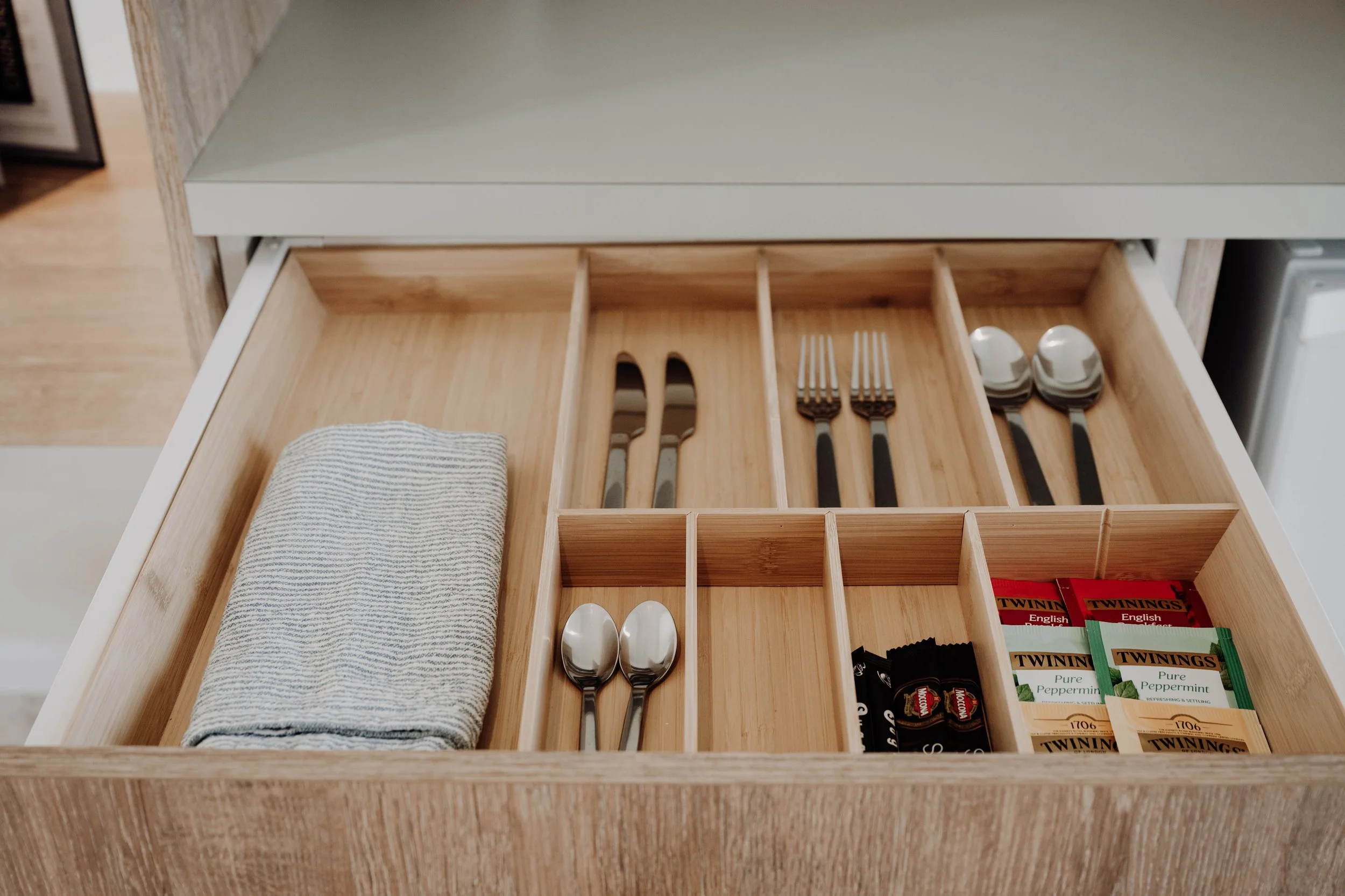 Open wooden kitchen drawer with organized compartments holding utensils, tea bags, a tea towel, and teaspoons.