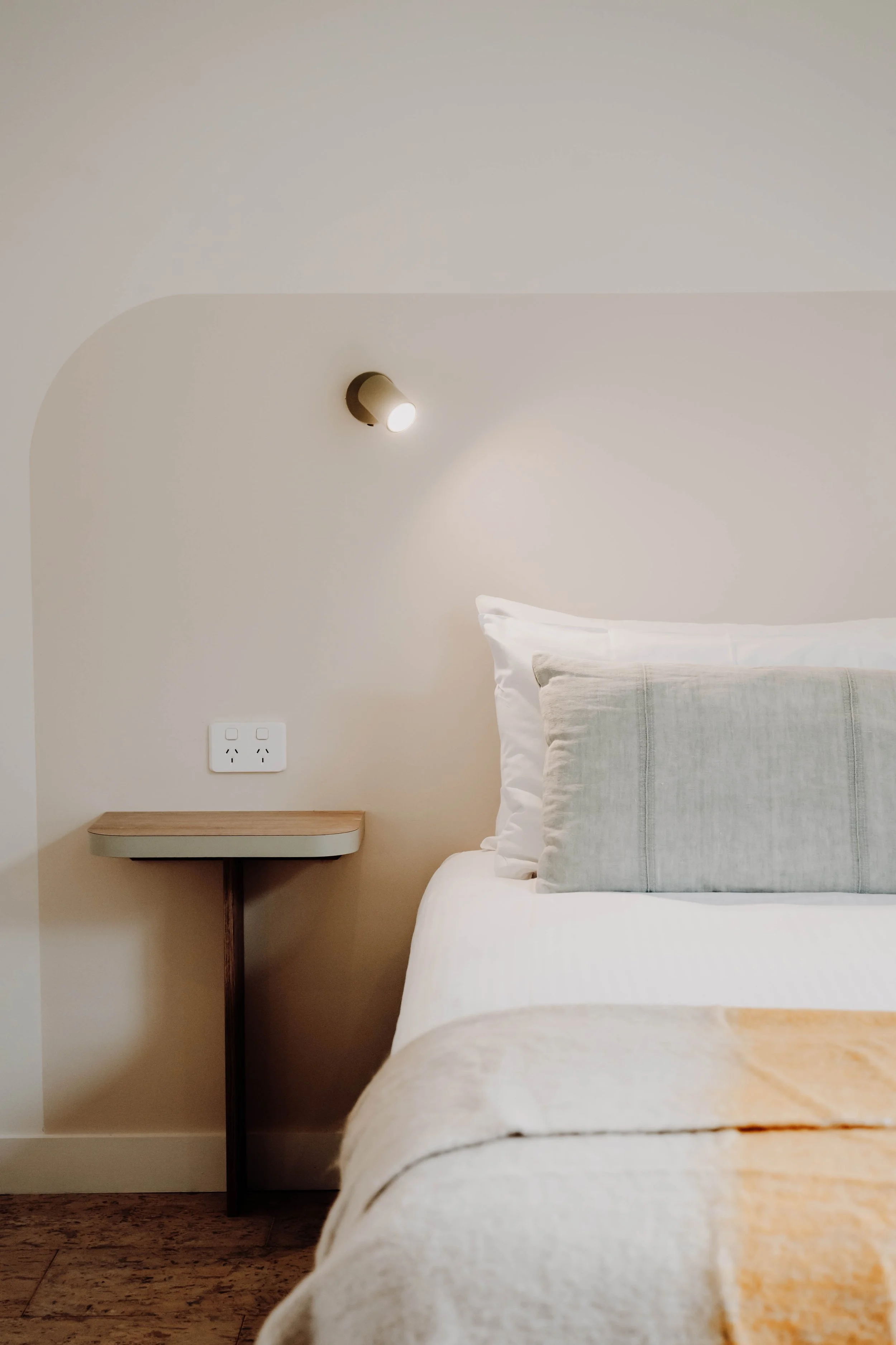 Close-up of a neatly made bed with light-colored pillows and blanket, a small bedside table, and a wall-mounted light fixture on a neutral wall.
