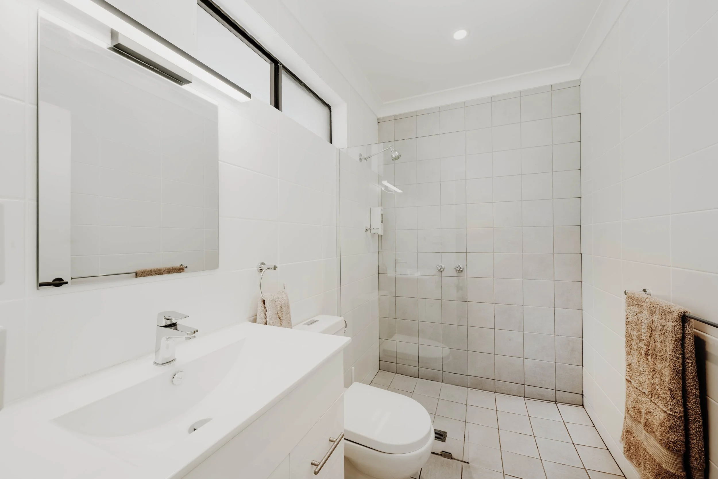 A clean, modern bathroom with white tiled walls and floor, a white sink with a faucet, a mirror above the sink, a toilet, and a glass shower enclosure with a showerhead.