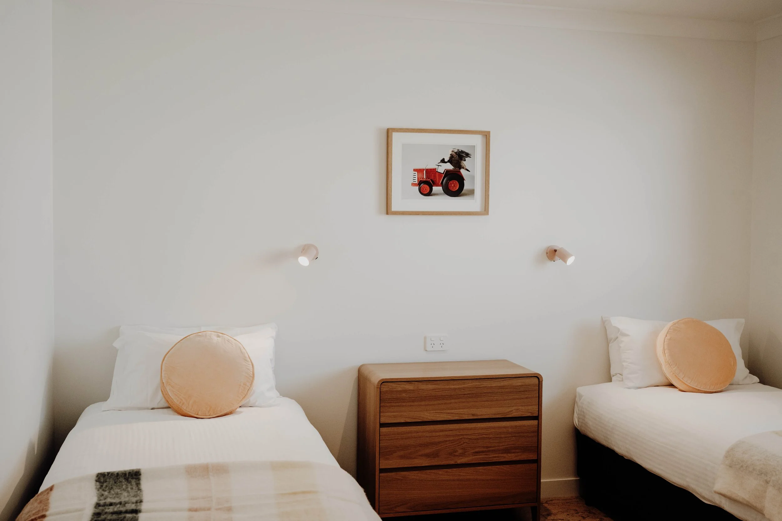 A bedroom with two single beds, each with white bedding and a round beige pillow, separated by a wooden nightstand. Above the beds, two wall-mounted reading lamps and a framed picture of a toy tractor with a bird on top are visible.