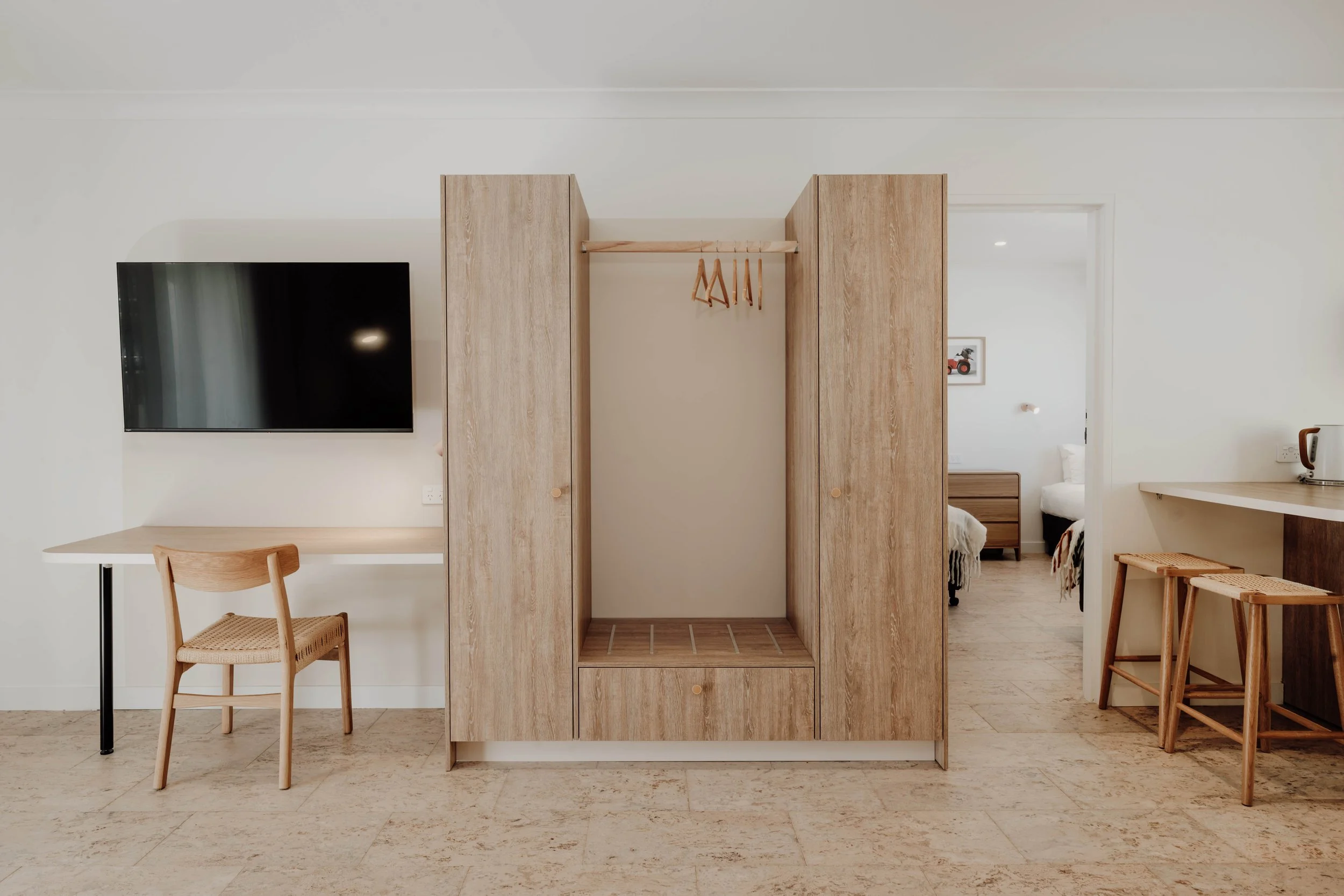 Modern hotel room with a built-in wooden wardrobe at center, a flat-screen TV on the wall to the left, a desk and chair underneath the TV, a partial view of a bedroom with a bed and dresser through an open doorway to the right, and two bar stools nex