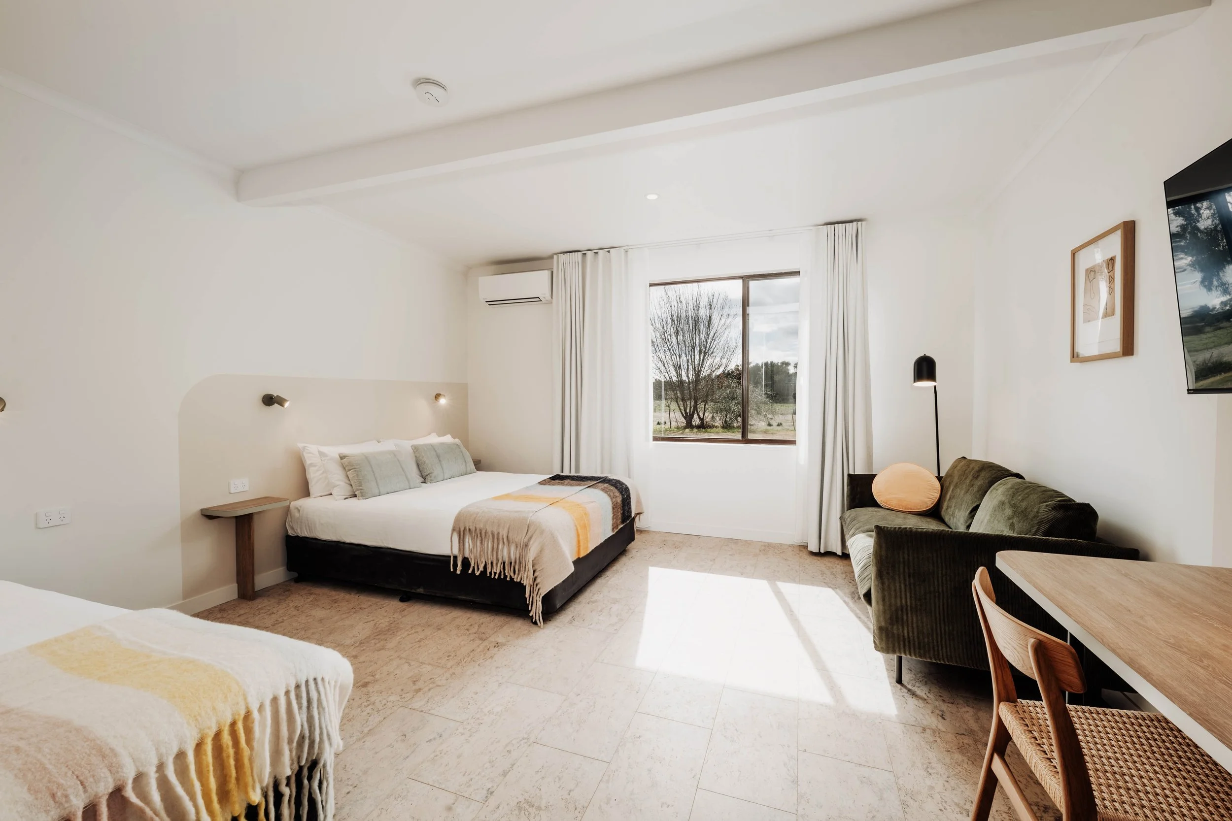 A bright hotel room with a large bed, small nightstands, a window with white curtains, a green sofa with a round pillow, a floor lamp, a wall-mounted TV, and a wooden dining table with chairs.
