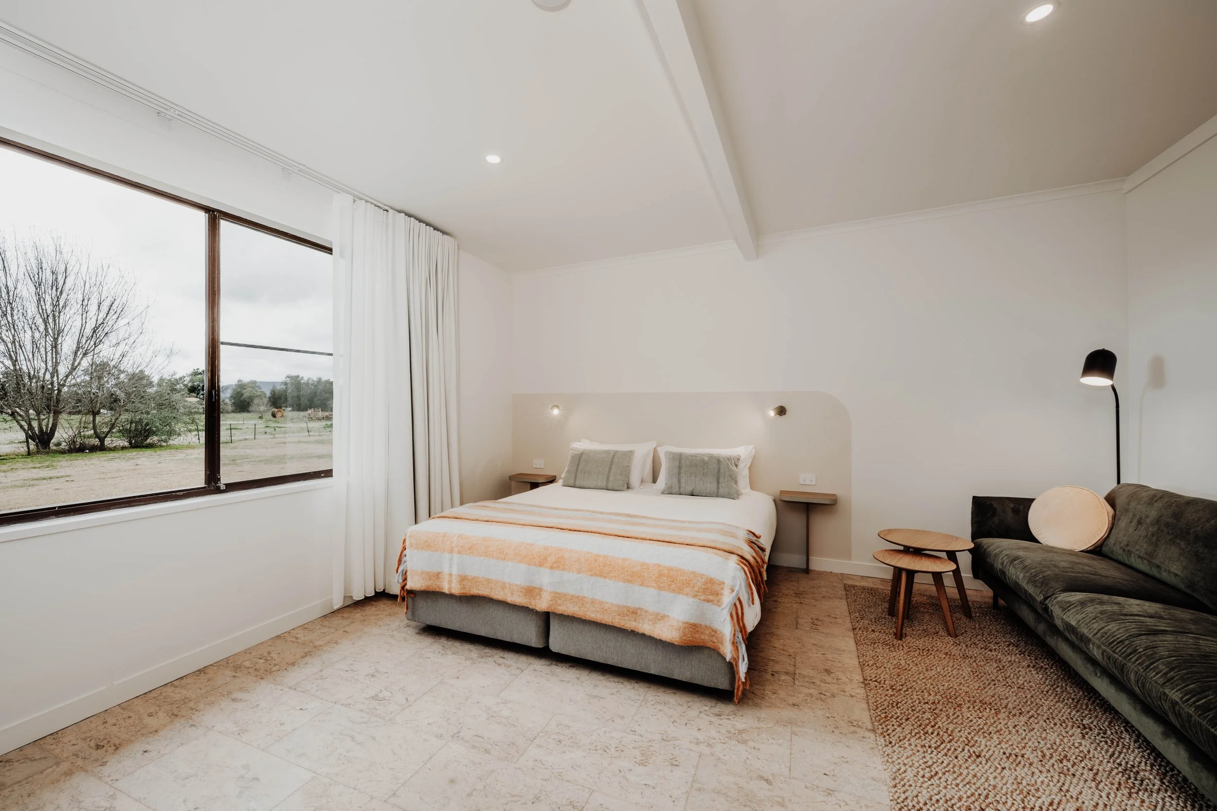 A bedroom featuring a large window with white curtains, a bed with white bedding and two green pillows, two side tables, a gray sofa with a round pillow, and a floor lamp with a black shade, with hardwood flooring and white walls.