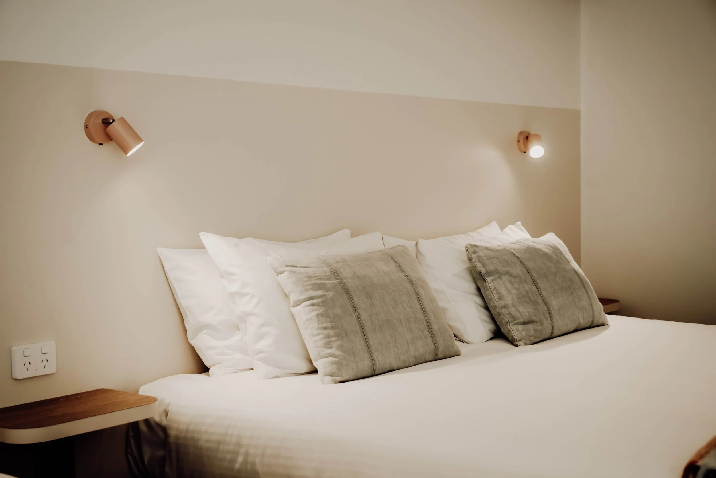 A bed with white sheets and four pillows, two beige and two white, in a softly lit bedroom with cream-colored walls and two wall-mounted bedside lamps.