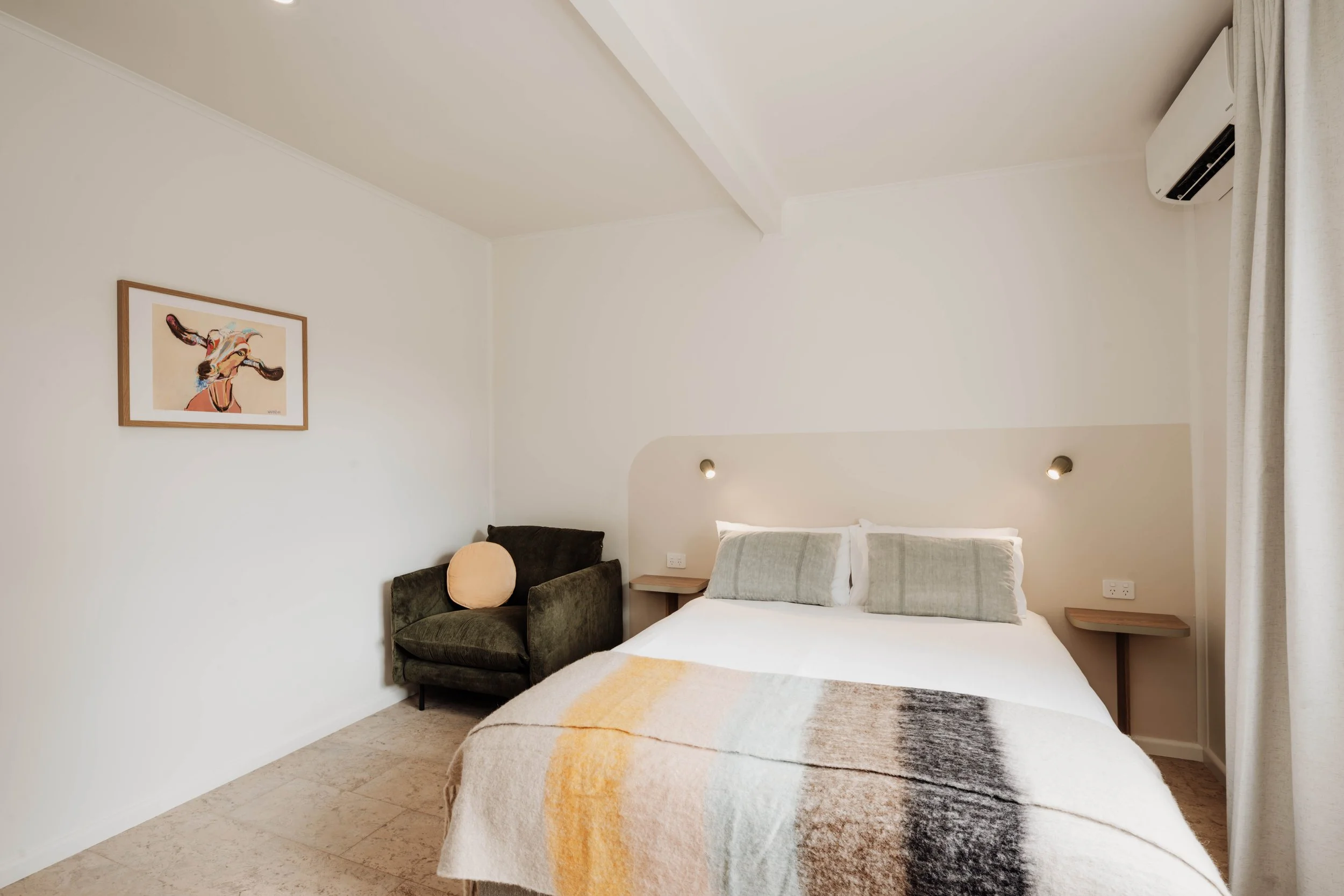 A bedroom with a bed, two pillows, and a blanket. There is an armchair with a round pillow and a framed painting of a cow on the wall. The room has a window with curtains and an air conditioning unit.