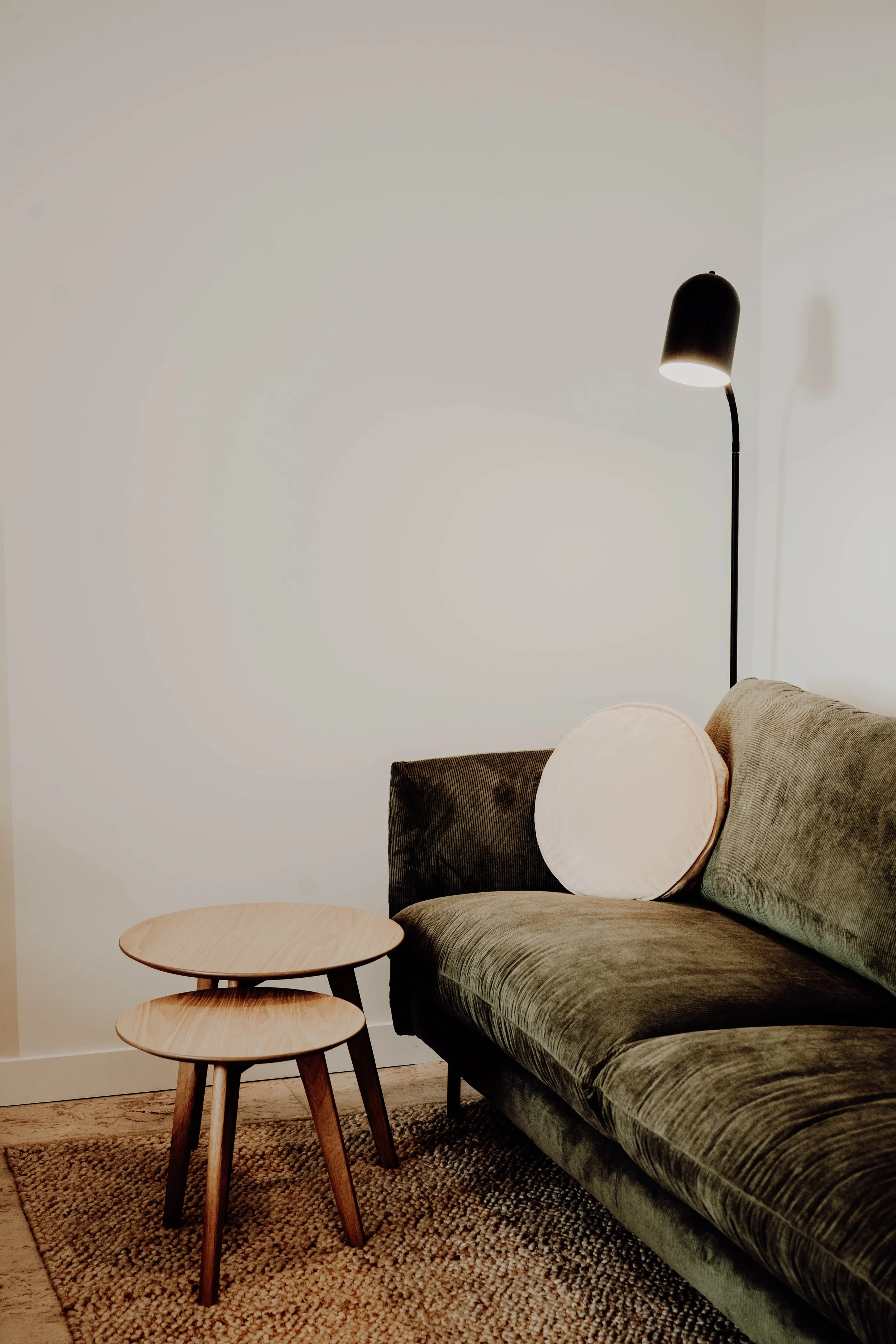 A cozy living room corner with a green velvet sofa, a round beige cushion, a pair of wooden nesting side tables, a textured area rug, and a tall black floor lamp with a curved neck illuminating the space.