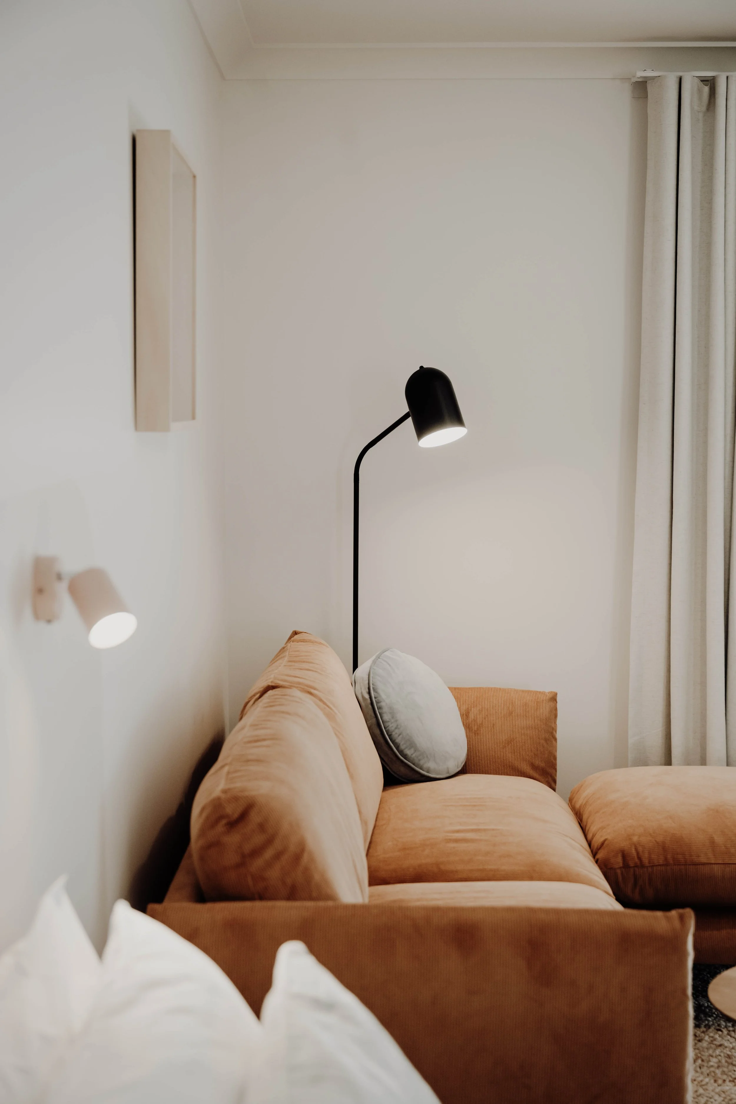 A cozy living room corner with a peach-colored couch, a round gray pillow, wall-mounted and standing lamps, and cream-colored curtains.