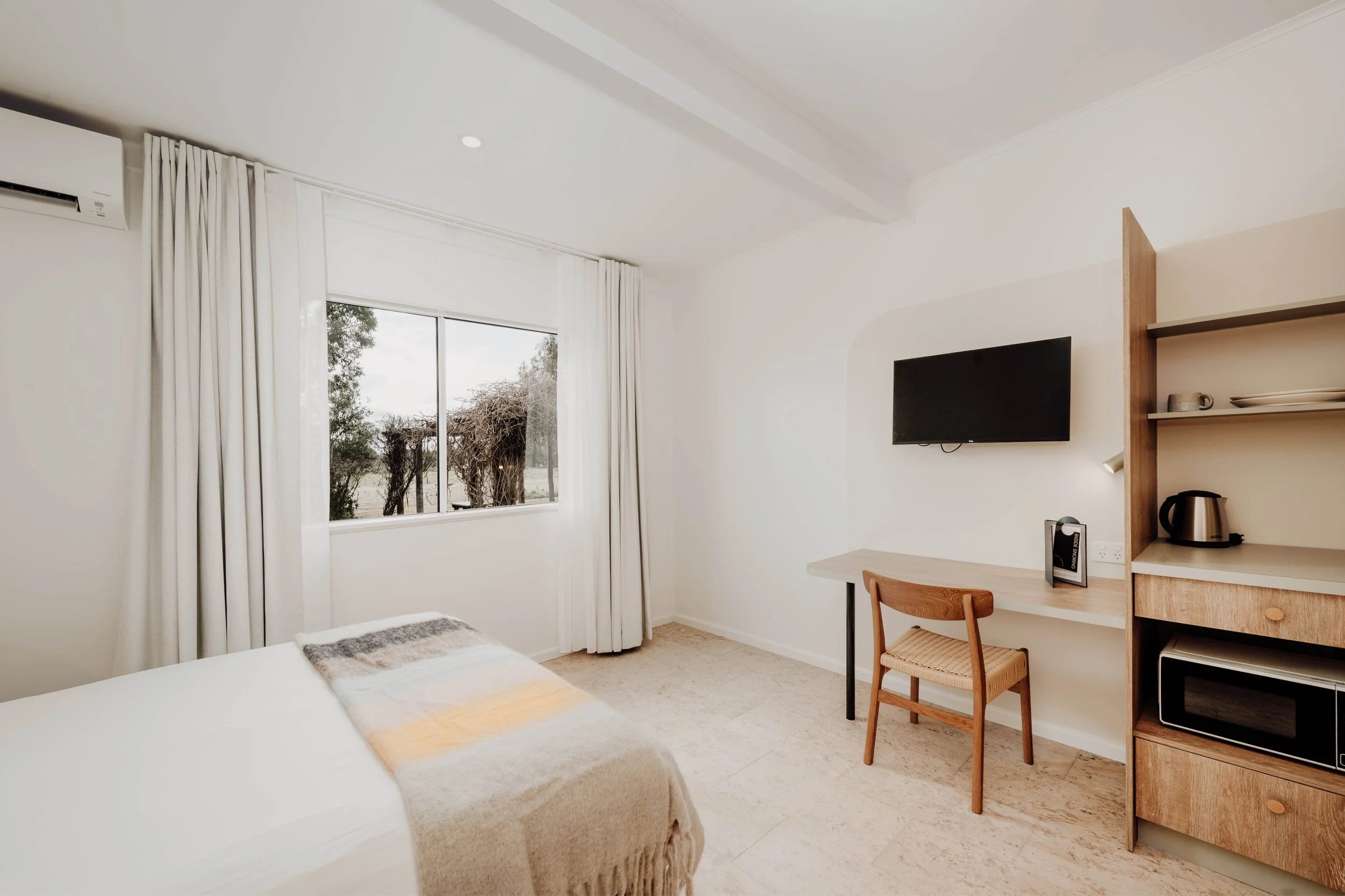 A minimalist hotel room with a bed, a window with white curtains, a wall-mounted TV, a wooden desk with a chair, and an open shelving unit with a kettle, microwave, and dishes.